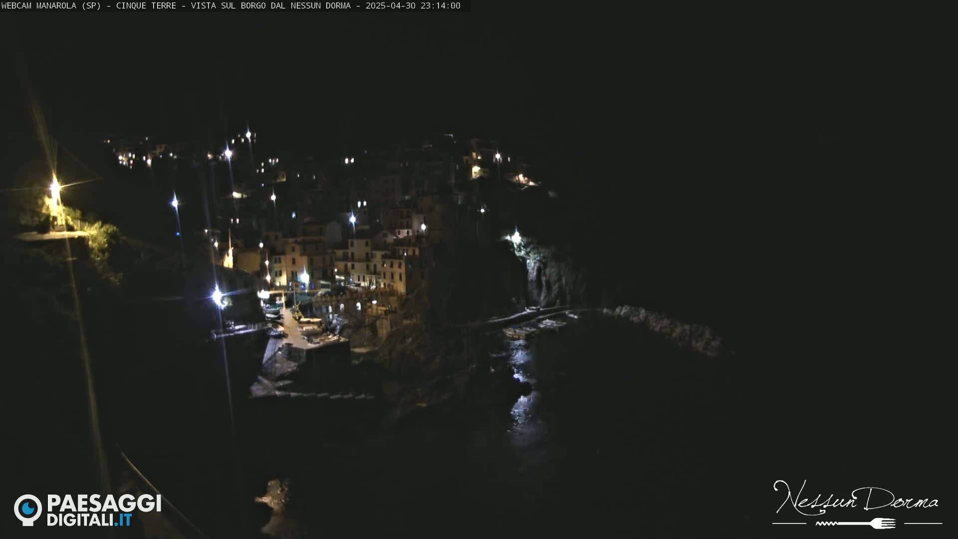 A nighttime view of a coastal town, illuminated by lights, with dark water visible below.