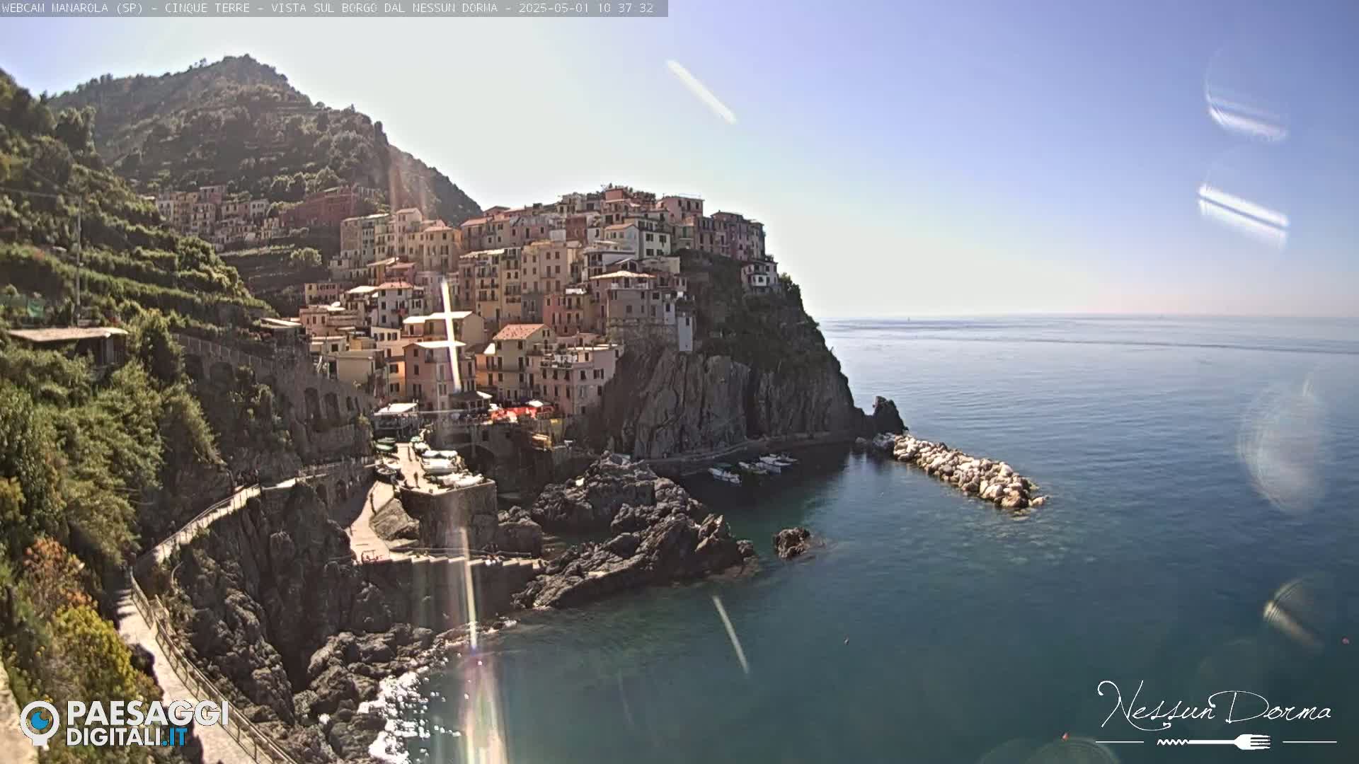A colorful village clings to a steep cliffside overlooking a calm, blue sea on a sunny day.