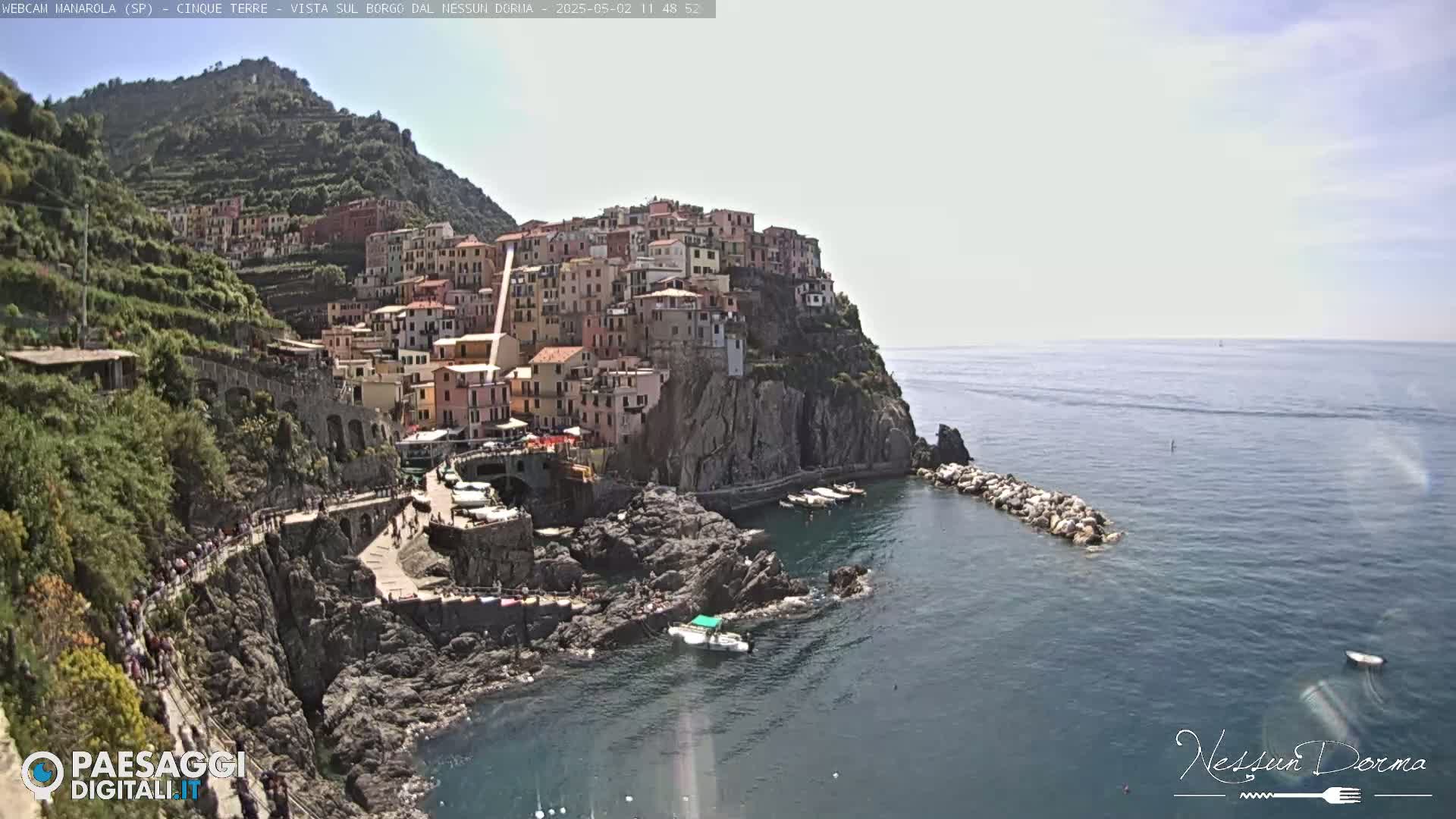 A colorful village clings to a steep, rocky cliffside overlooking a calm, blue sea on a mostly clear day.