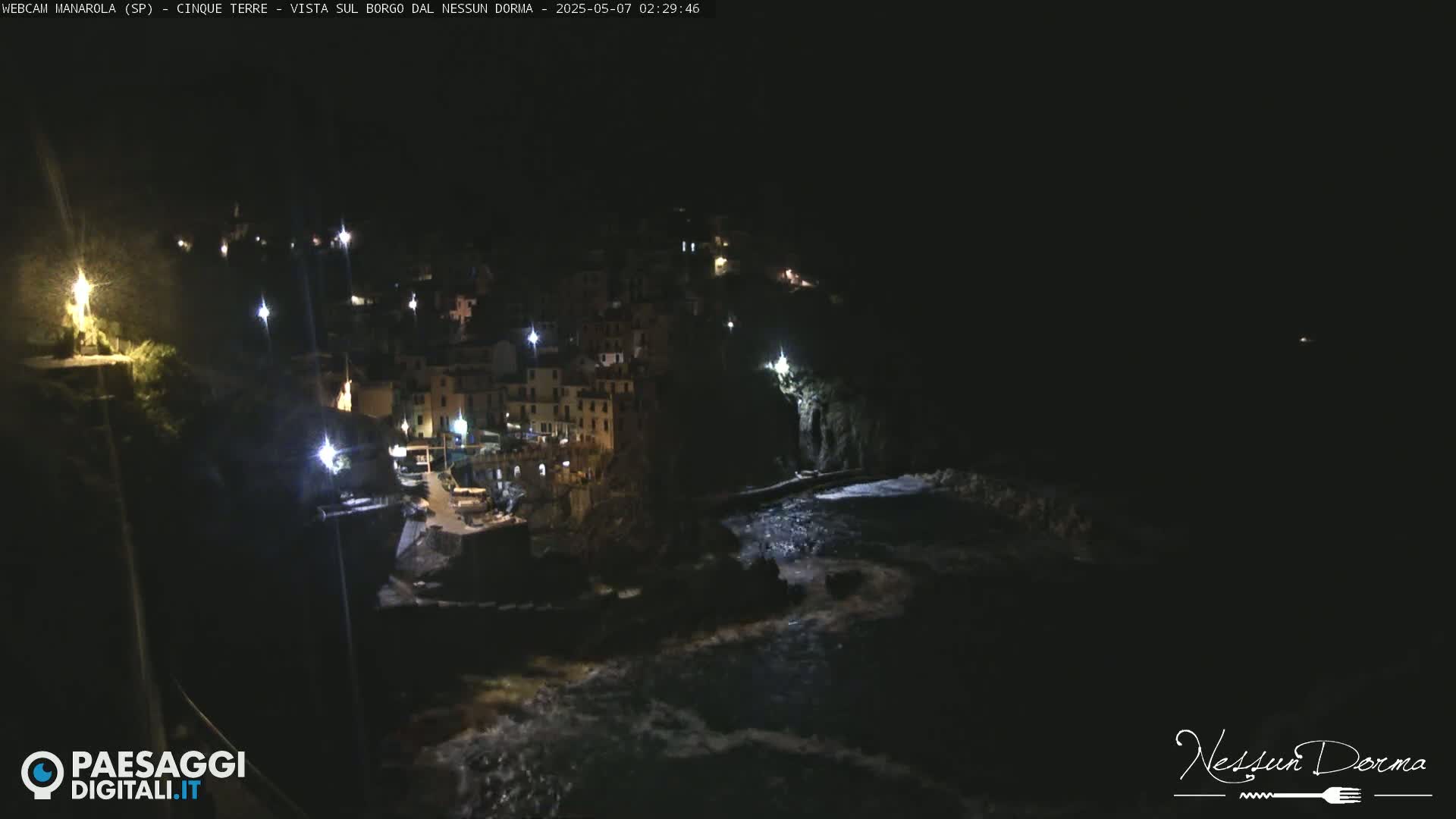 A nighttime coastal village with buildings illuminated by lights is partially visible, overlooking a dark, wavy ocean.