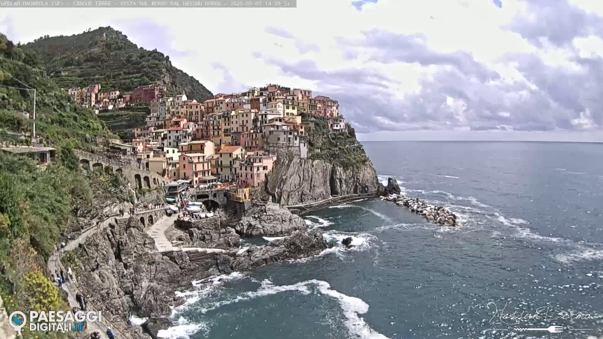 A colorful village clings to a steep cliffside overlooking a partly cloudy ocean.