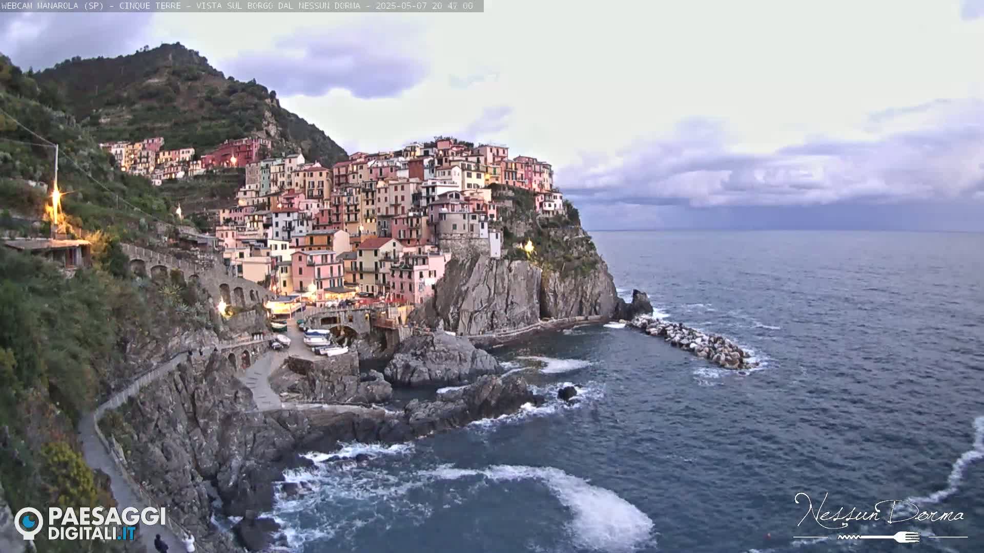 A colorful village clings to a steep cliffside overlooking a dark, wavy sea under a partly cloudy sky.