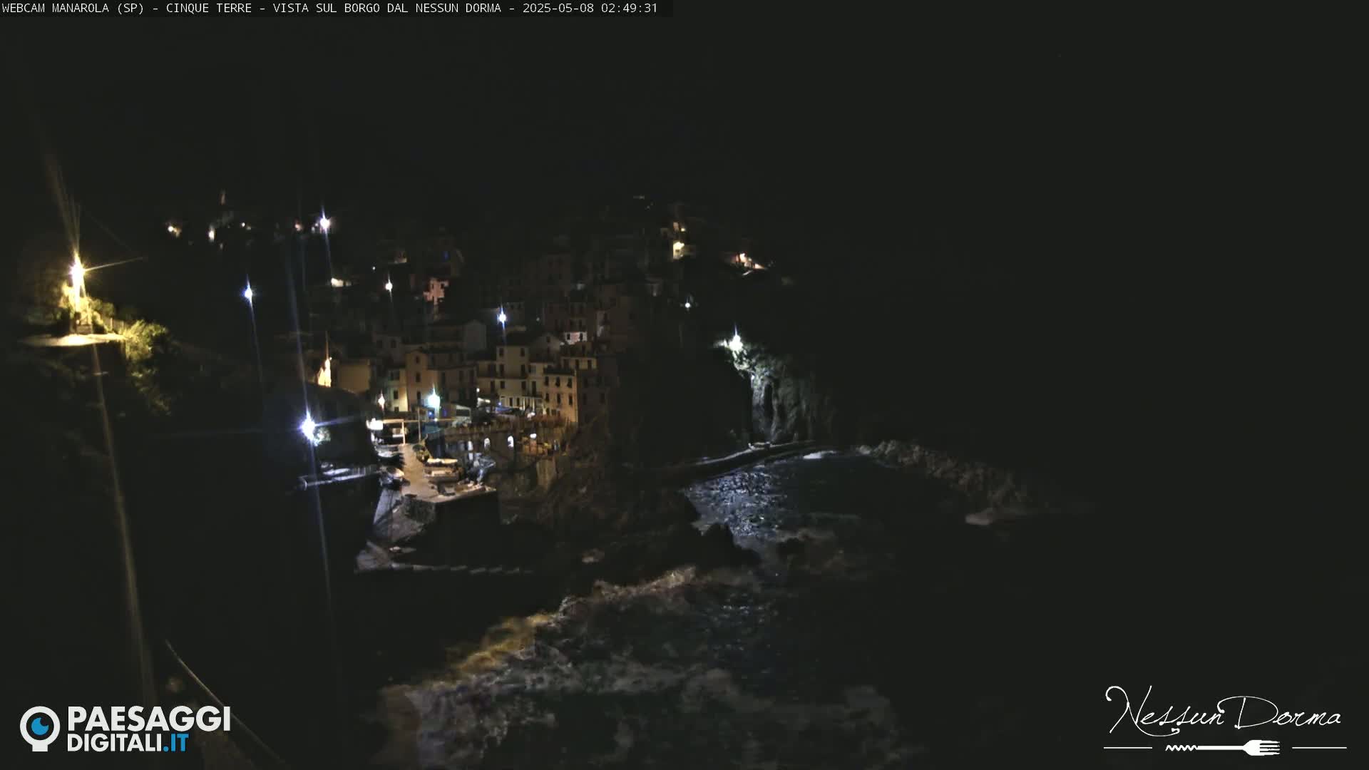 A nighttime, coastal village is illuminated by scattered lights, with dark, choppy water visible below.