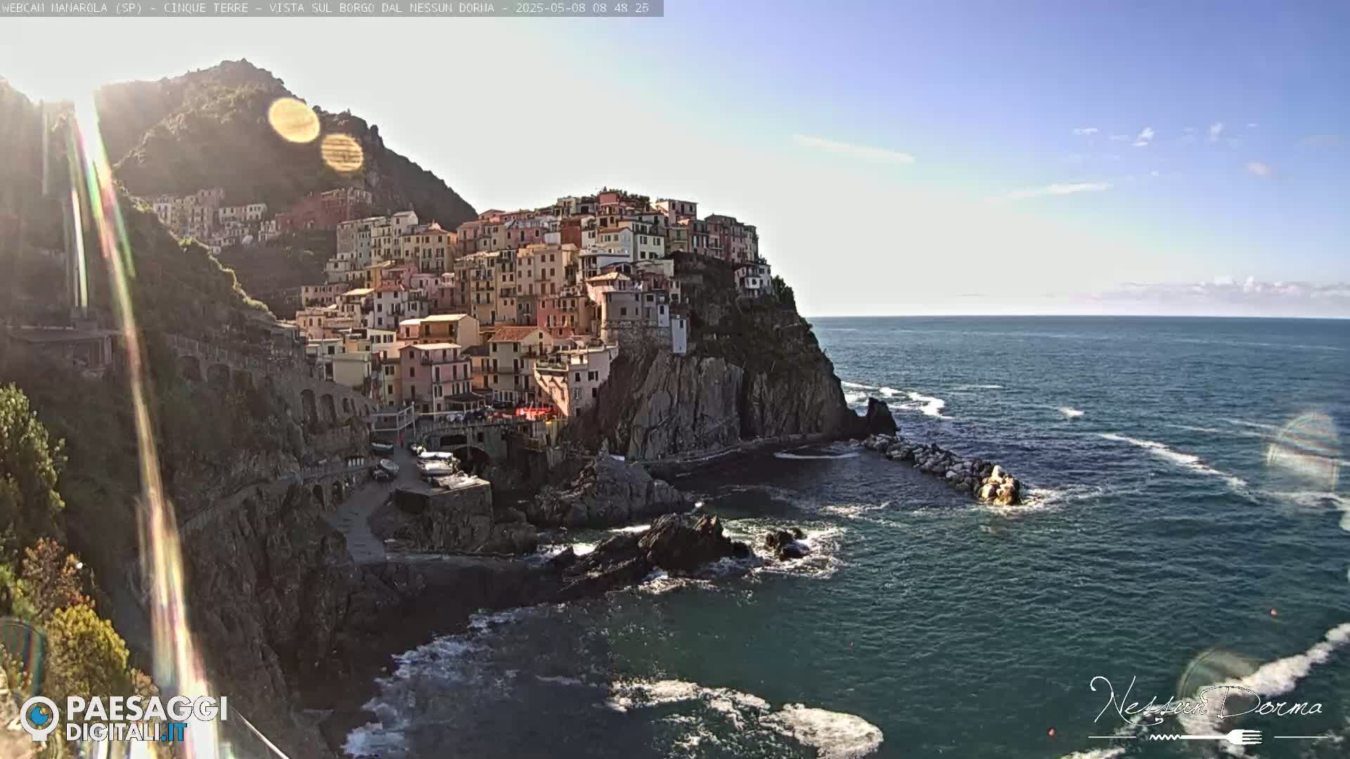 A picturesque coastal village clings to a cliffside overlooking a calm sea under a mostly sunny sky.