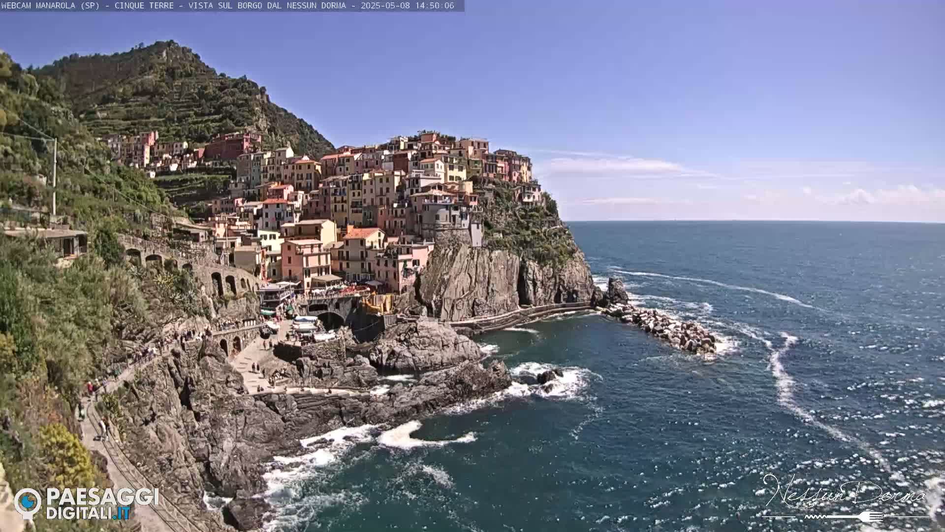 A colorful village clings to a steep cliffside overlooking a blue ocean on a sunny day.