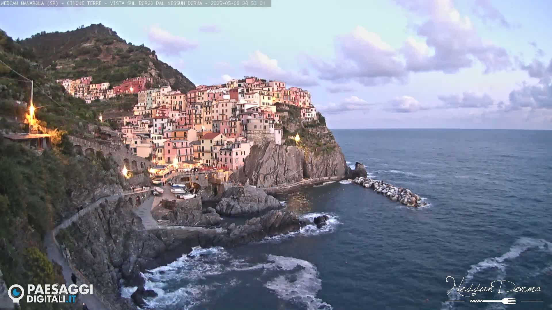 A colorful village clings to a steep cliffside overlooking a dark blue sea under a partly cloudy sky.