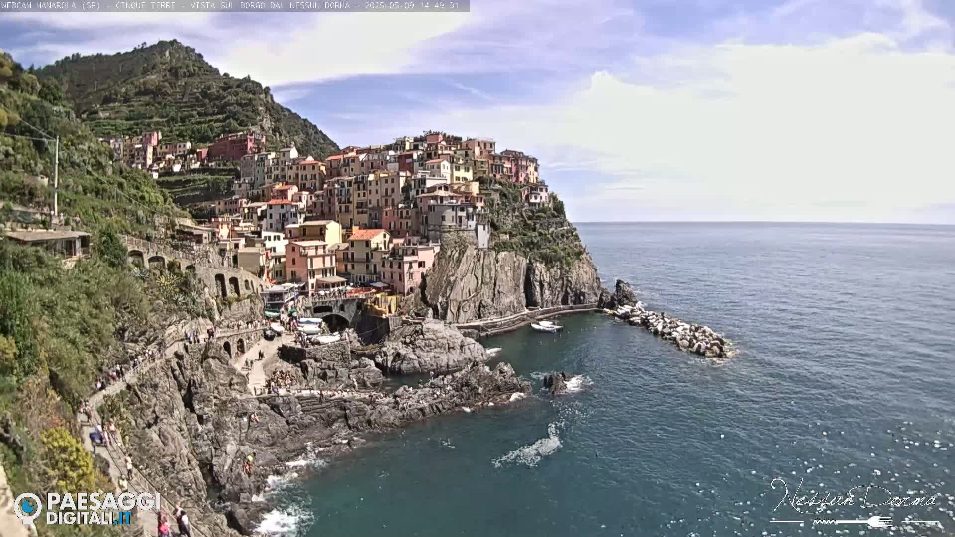 A colorful village clings to a steep cliffside overlooking a calm, blue sea under a mostly sunny sky.