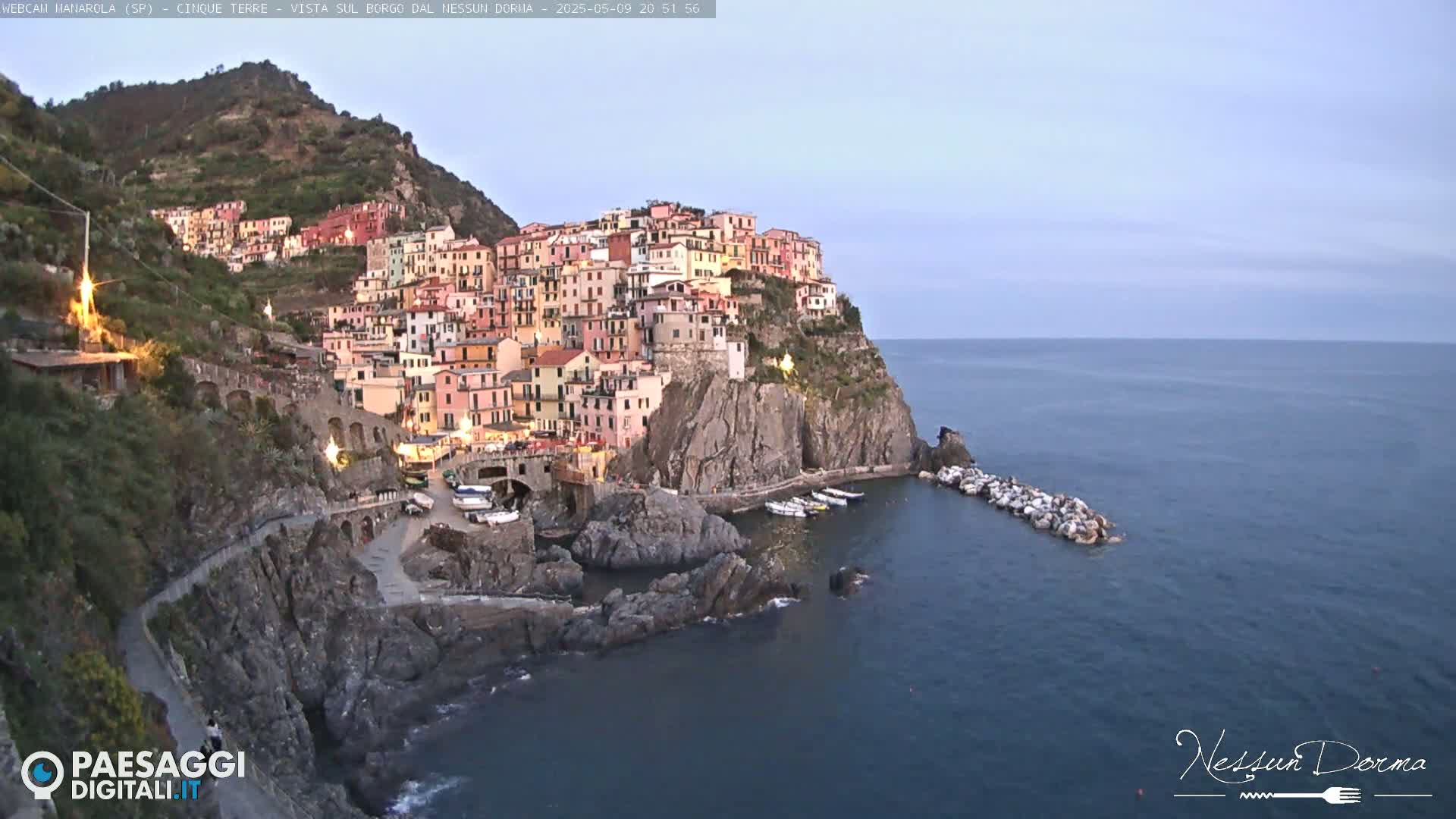 A colorful village clings to a cliffside overlooking a calm, dark-blue sea at dusk.