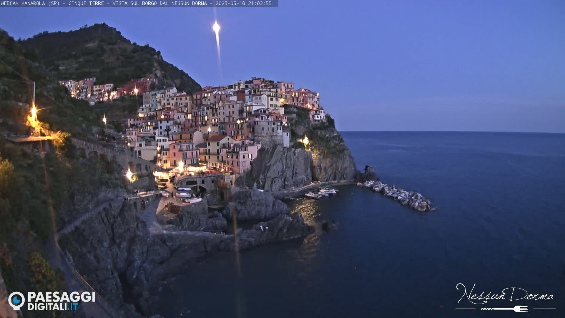 A brightly lit coastal town nestled on a cliffside overlooking a dark blue sea under a twilight sky.