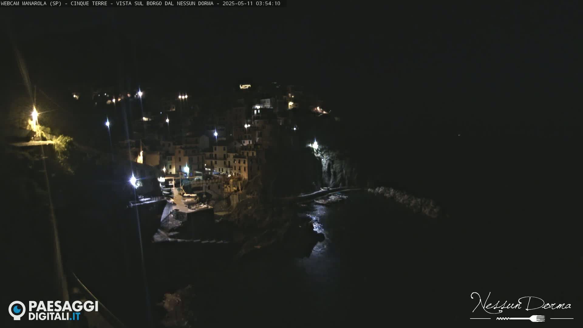 A nighttime aerial view shows a coastal town illuminated by lights, nestled against a dark hillside and partially surrounded by water.