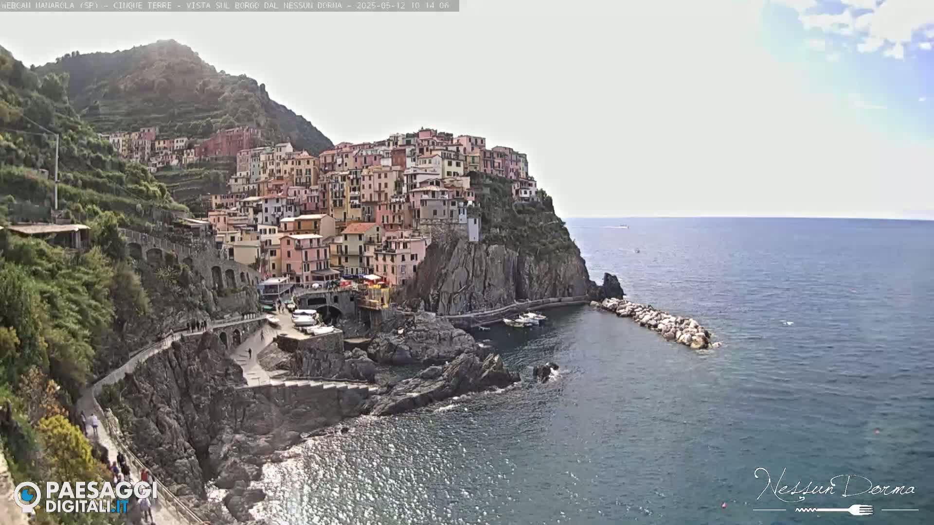 A colorful village clings to a steep cliffside overlooking a calm sea under a partly cloudy sky.
