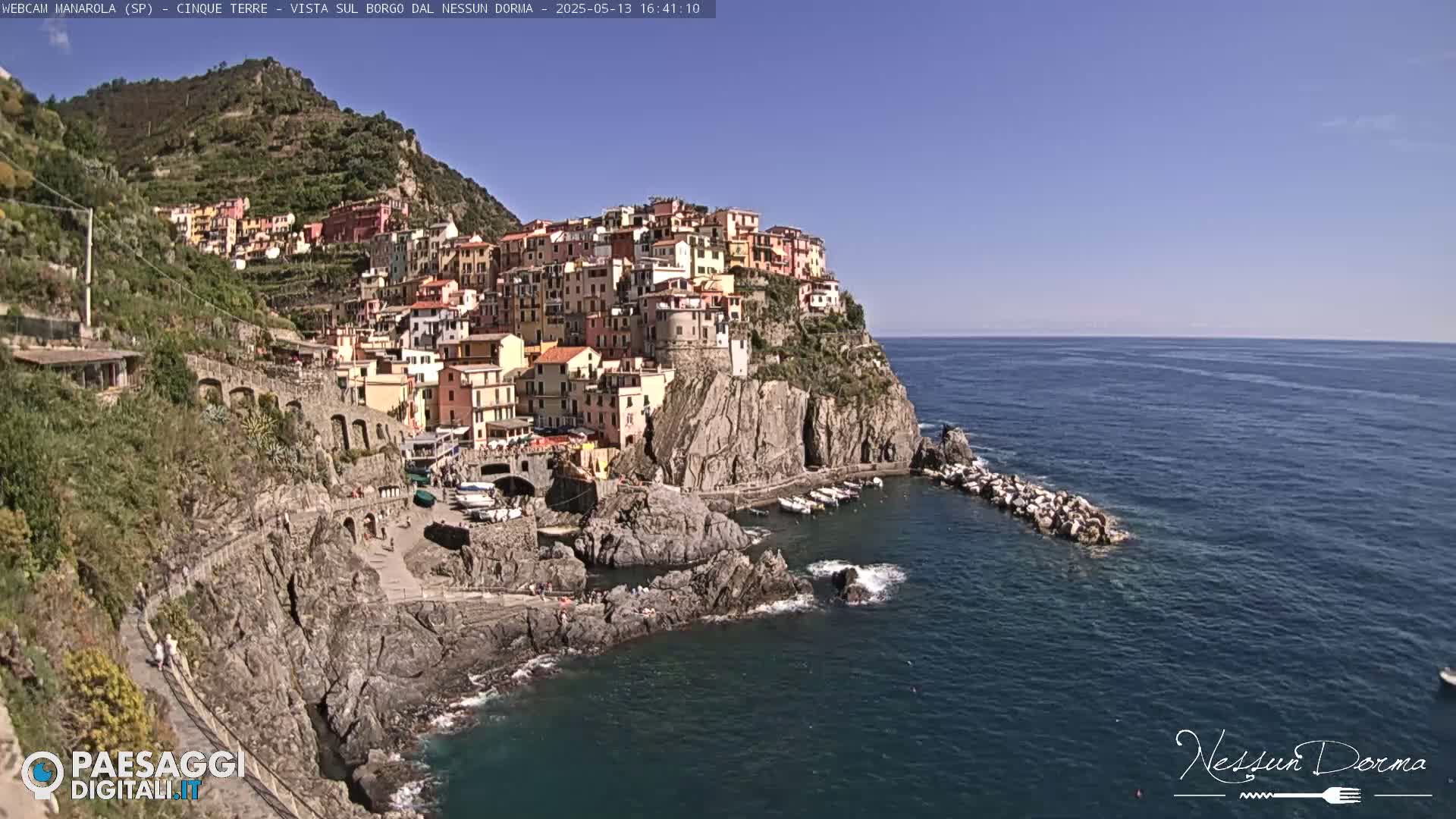 A colorful village clings to a rocky cliffside overlooking a calm blue sea under a clear sky.