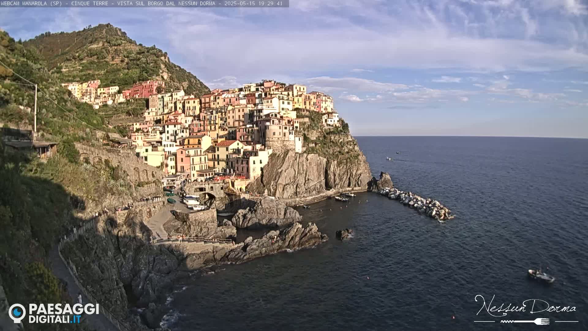 A colorful village clings to a steep cliffside overlooking a calm, blue sea under a mostly clear sky.