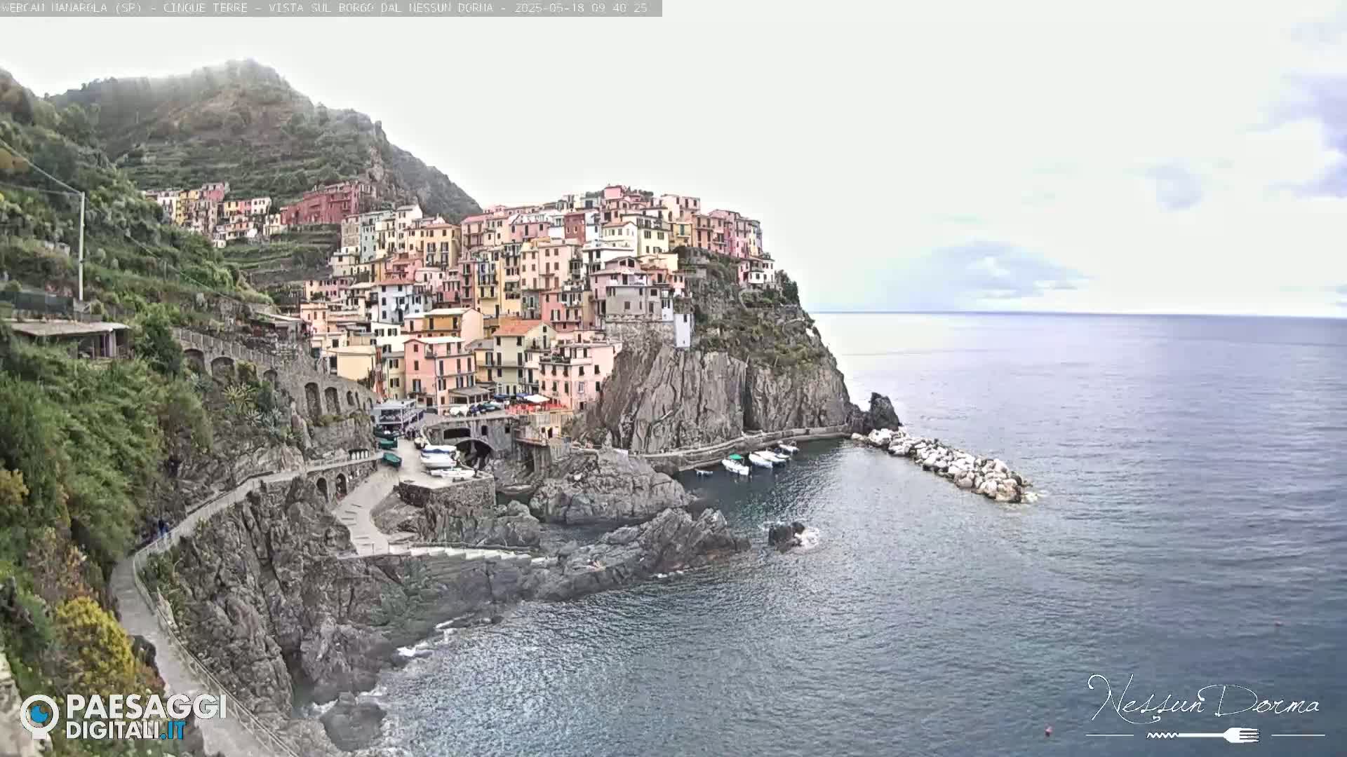 A colorful village clings to a steep cliffside overlooking a calm sea under a mostly cloudy sky.