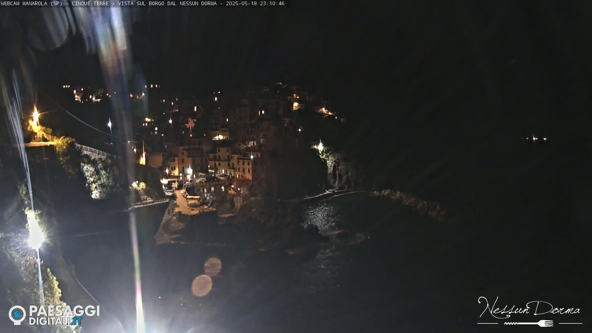 A nighttime view of a coastal town illuminated by lights, nestled against a dark hillside overlooking the sea.
