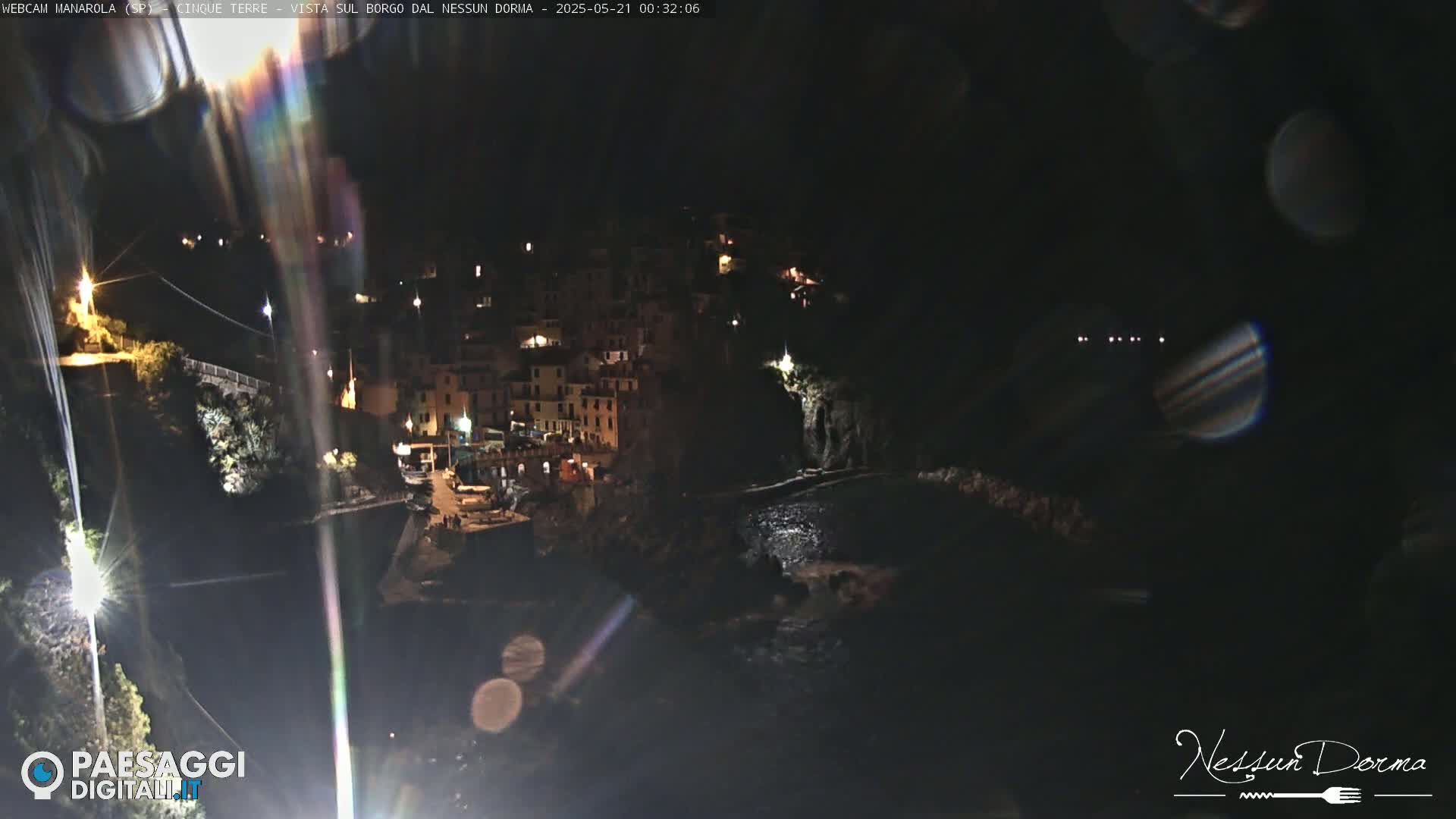 A nighttime, coastal village with brightly lit buildings clings to a dark cliffside, partially obscured by lens flares.