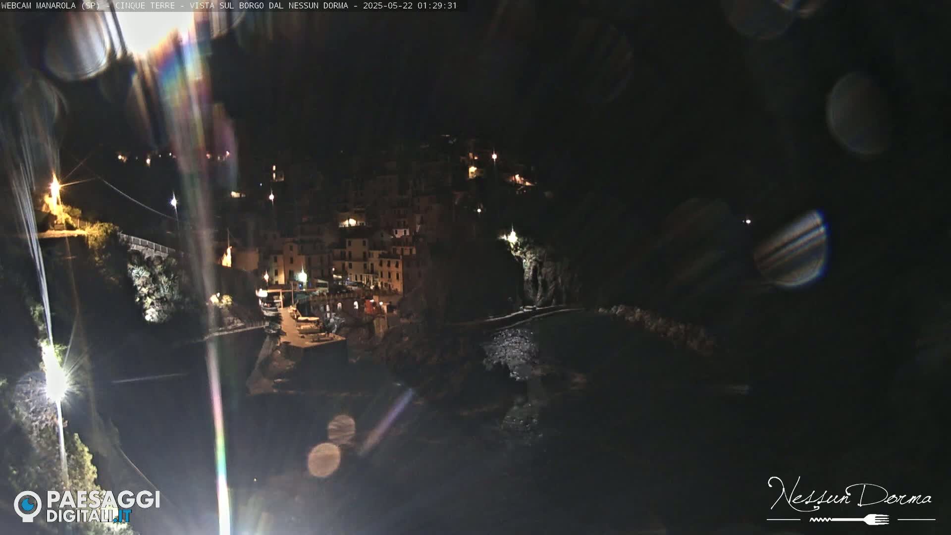 A nighttime, coastal village with buildings illuminated, nestled between dark cliffs and a calm body of water.