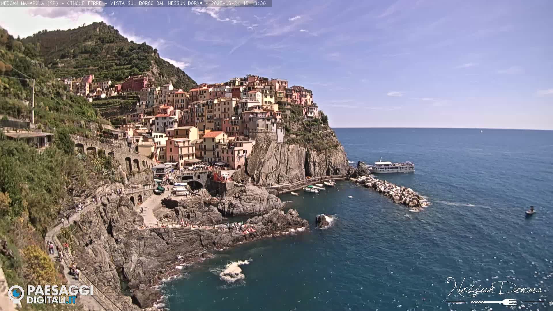 A colorful village clings to a rocky cliffside overlooking a calm blue sea, under a mostly sunny sky.