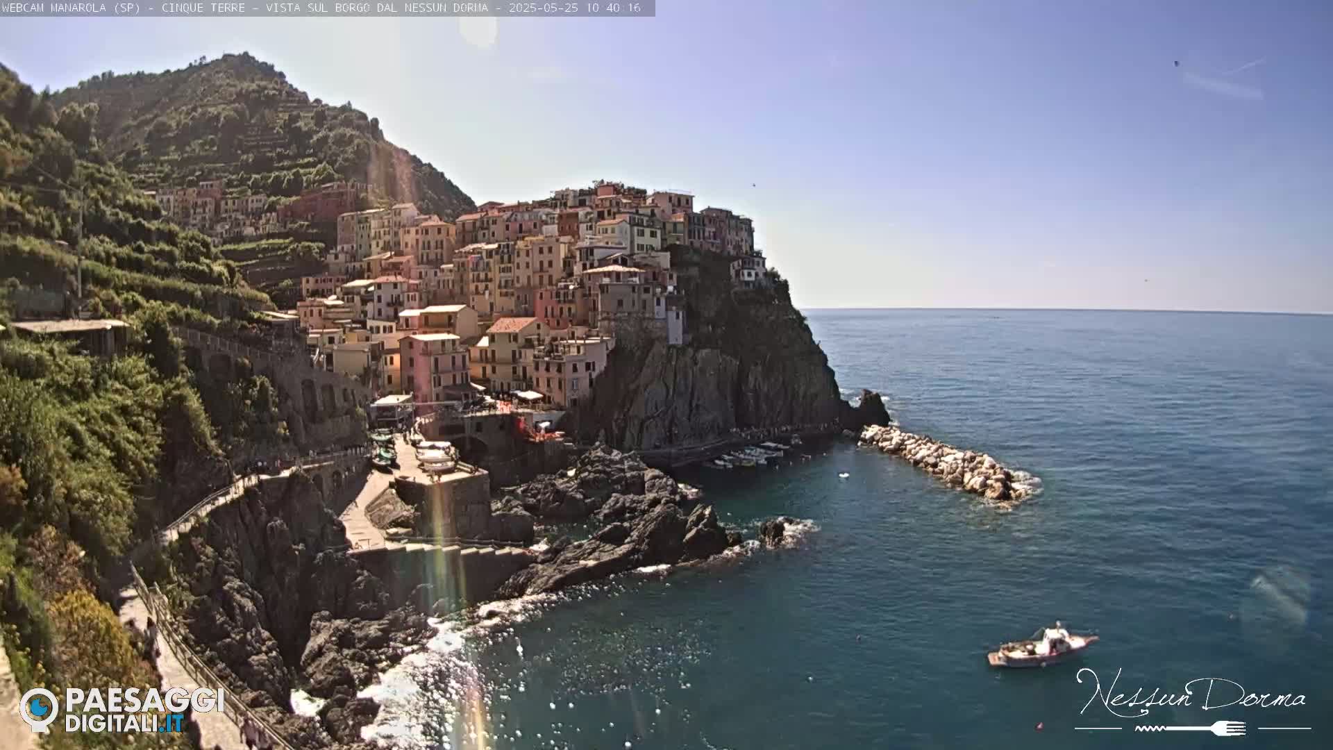 A picturesque coastal town, built on a steep cliffside, is shown on a sunny day with a calm sea and a small boat visible in the distance.