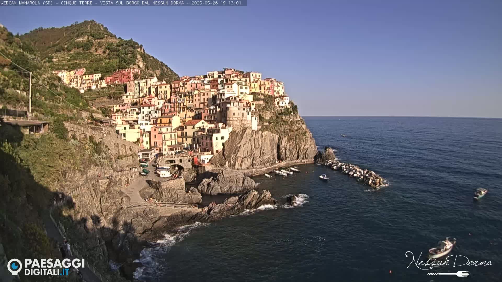 A colorful village clings to a steep cliffside overlooking a calm, blue sea dotted with small boats under a clear sky.