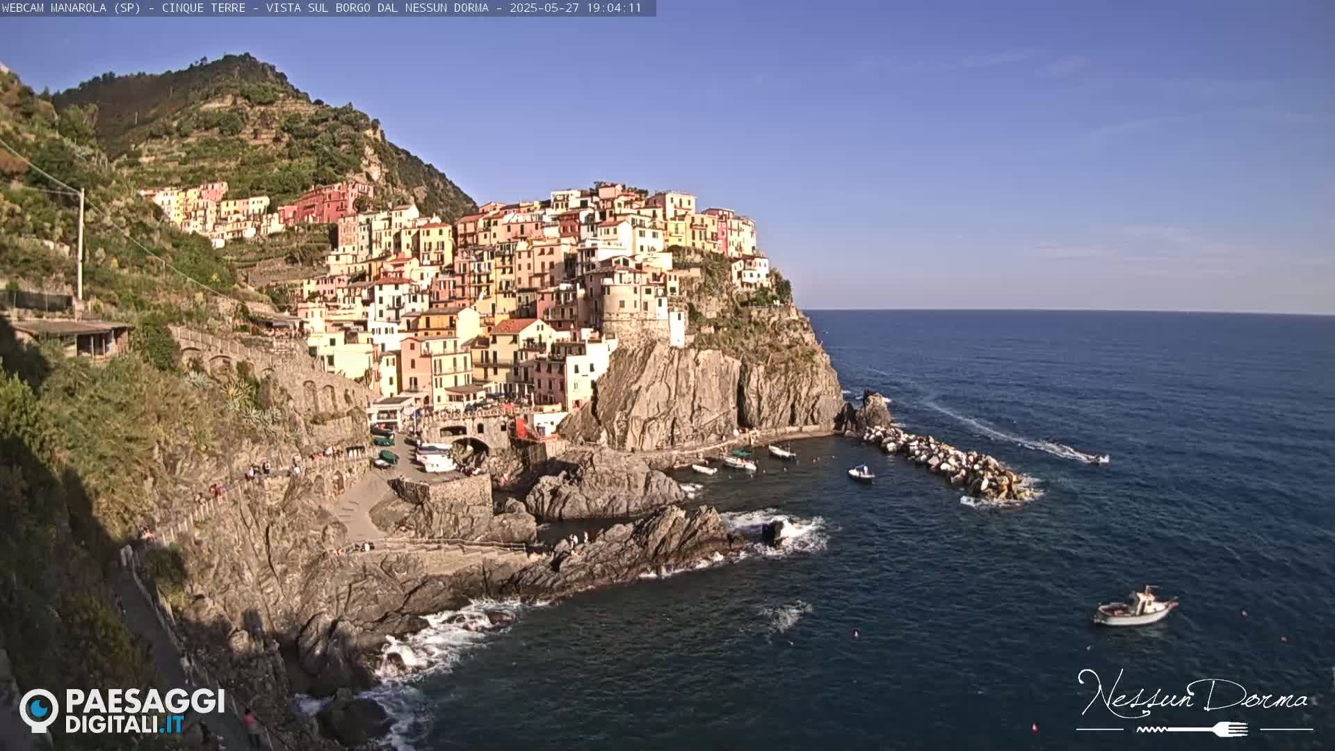 A colorful village clings to a steep cliffside overlooking a calm sea on a sunny day.