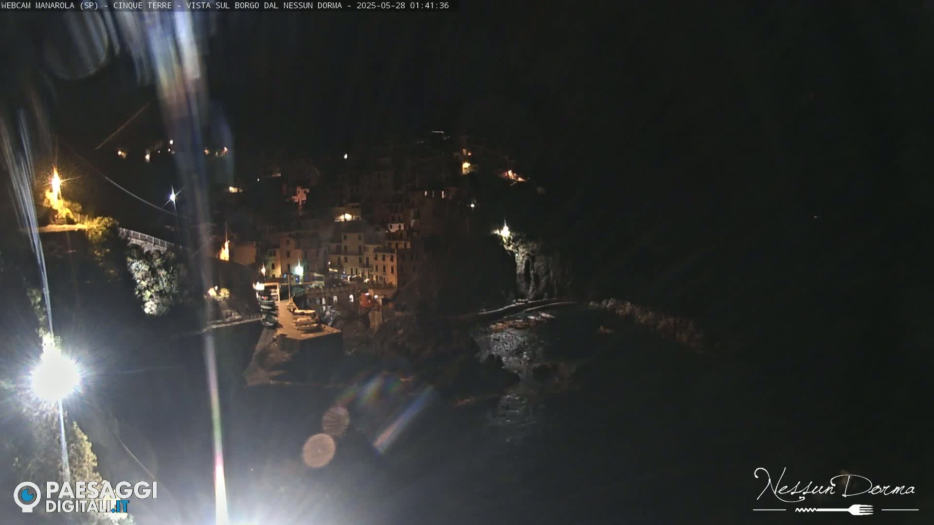 A nighttime, long shot of a coastal town with buildings lit up along a cliffside overlooking the dark ocean.