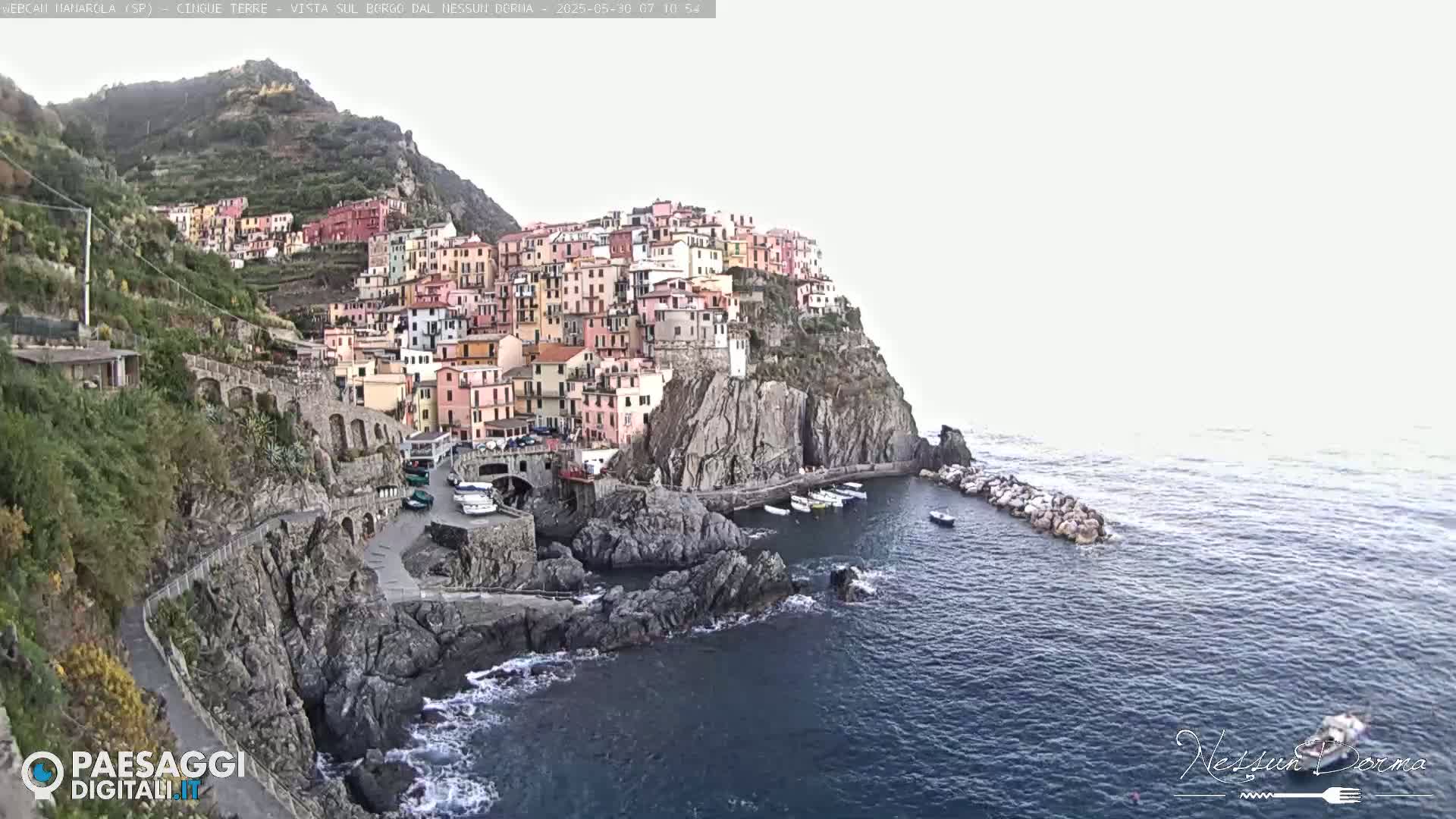 A colorful village clings to a steep cliffside overlooking a calm sea under a bright, overcast sky.