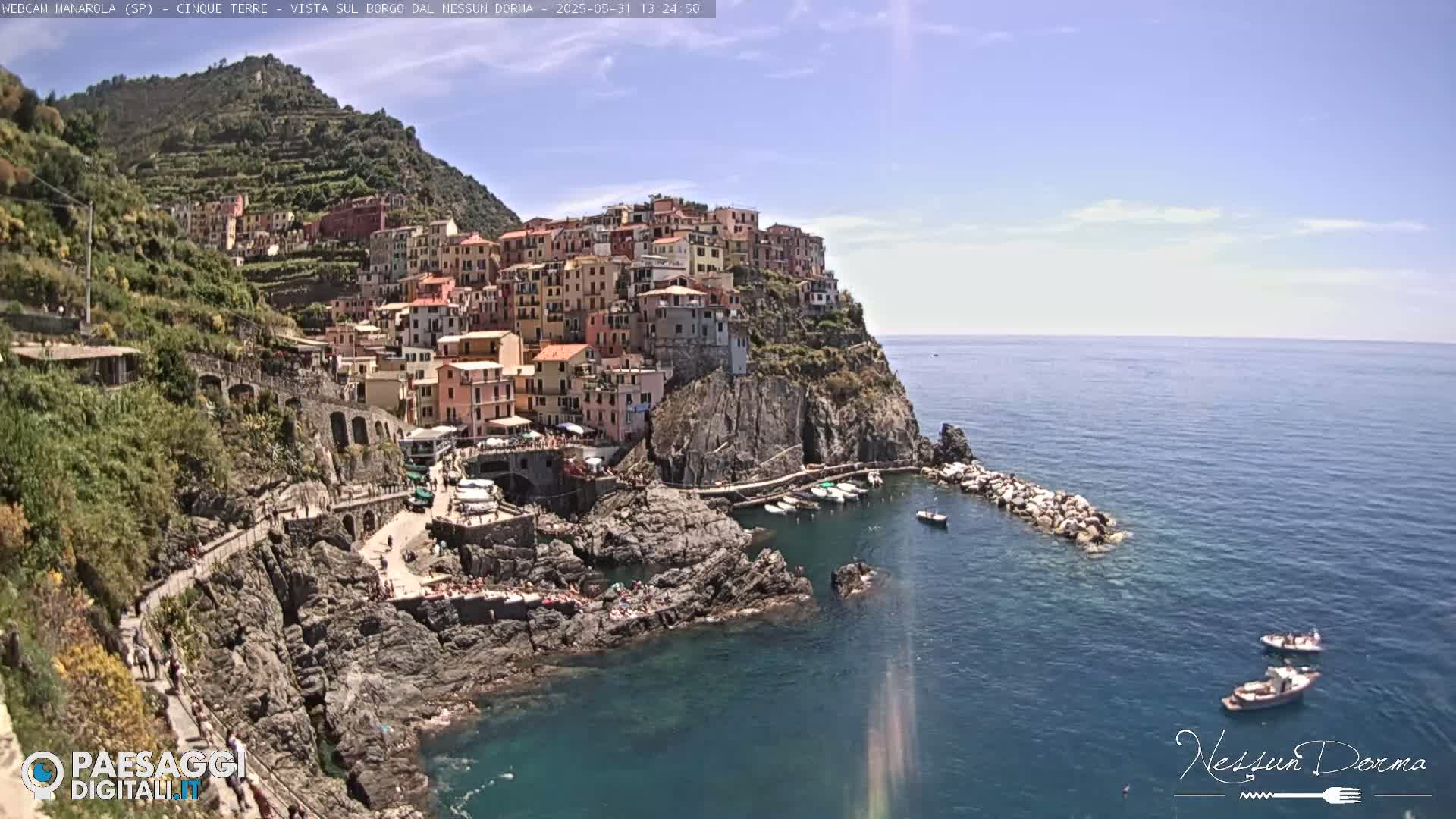 A colorful village clings to a rocky cliffside overlooking a calm, blue sea on a sunny day.