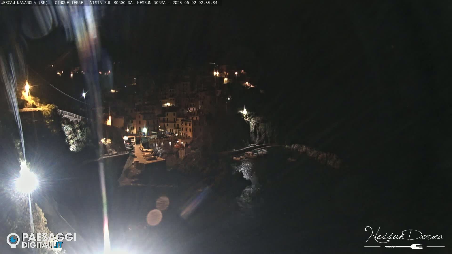 A nighttime, coastal village with buildings illuminated along a dark cliffside overlooking a calm body of water.