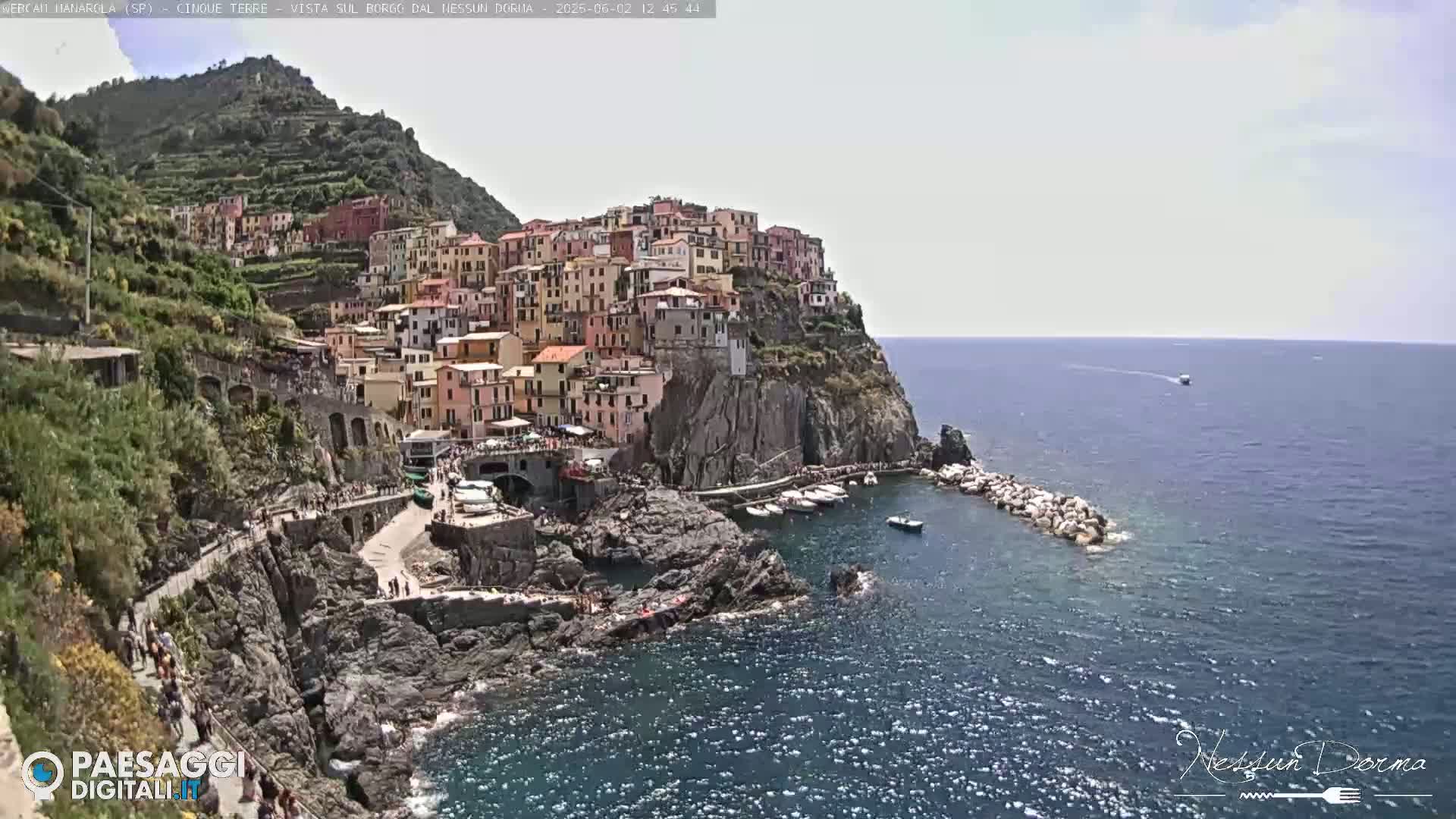 A colorful village clings to a rocky cliffside overlooking a calm sea on a partly sunny day.