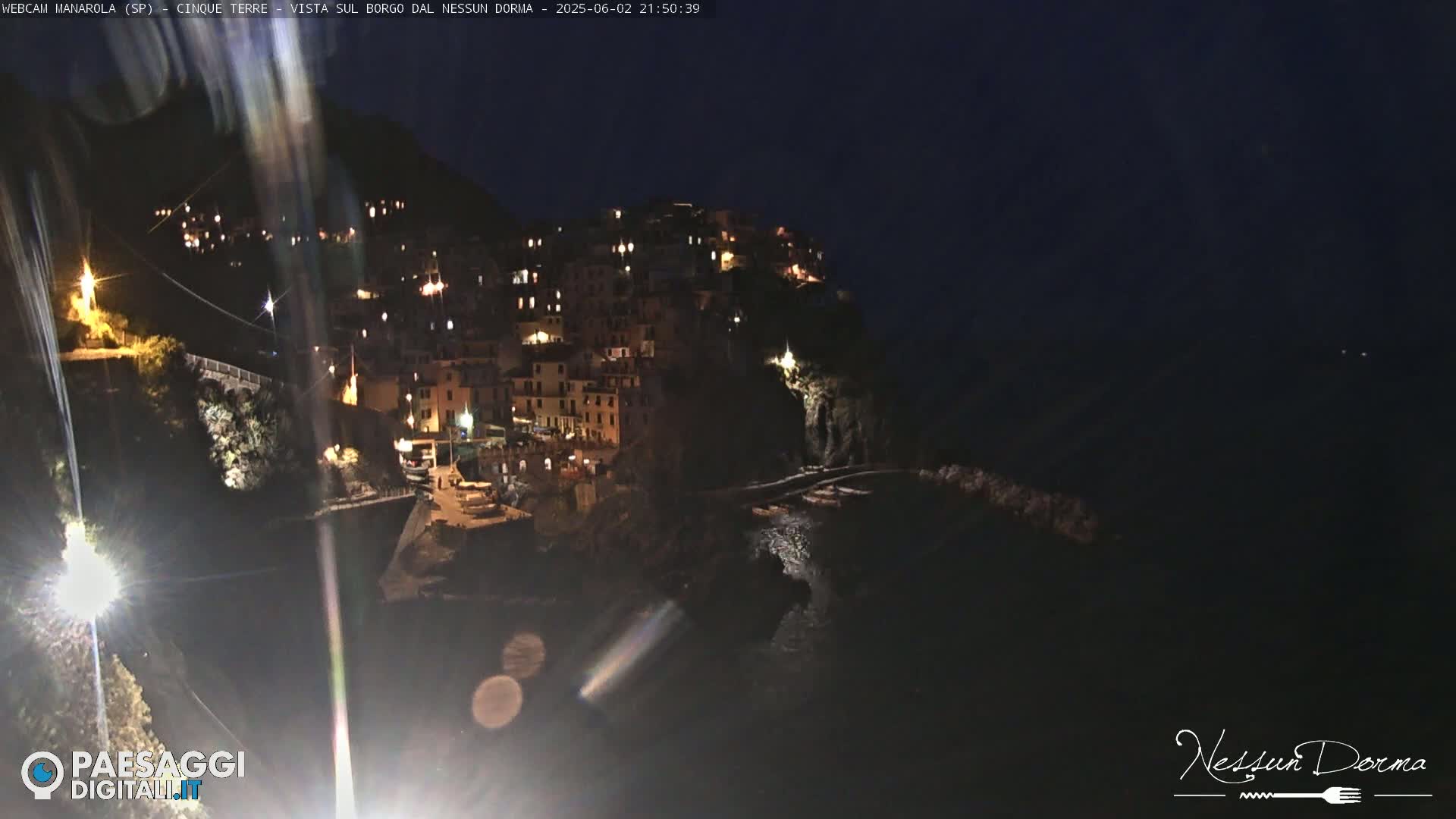 A nighttime view of a village nestled on a cliffside overlooking a dark body of water.