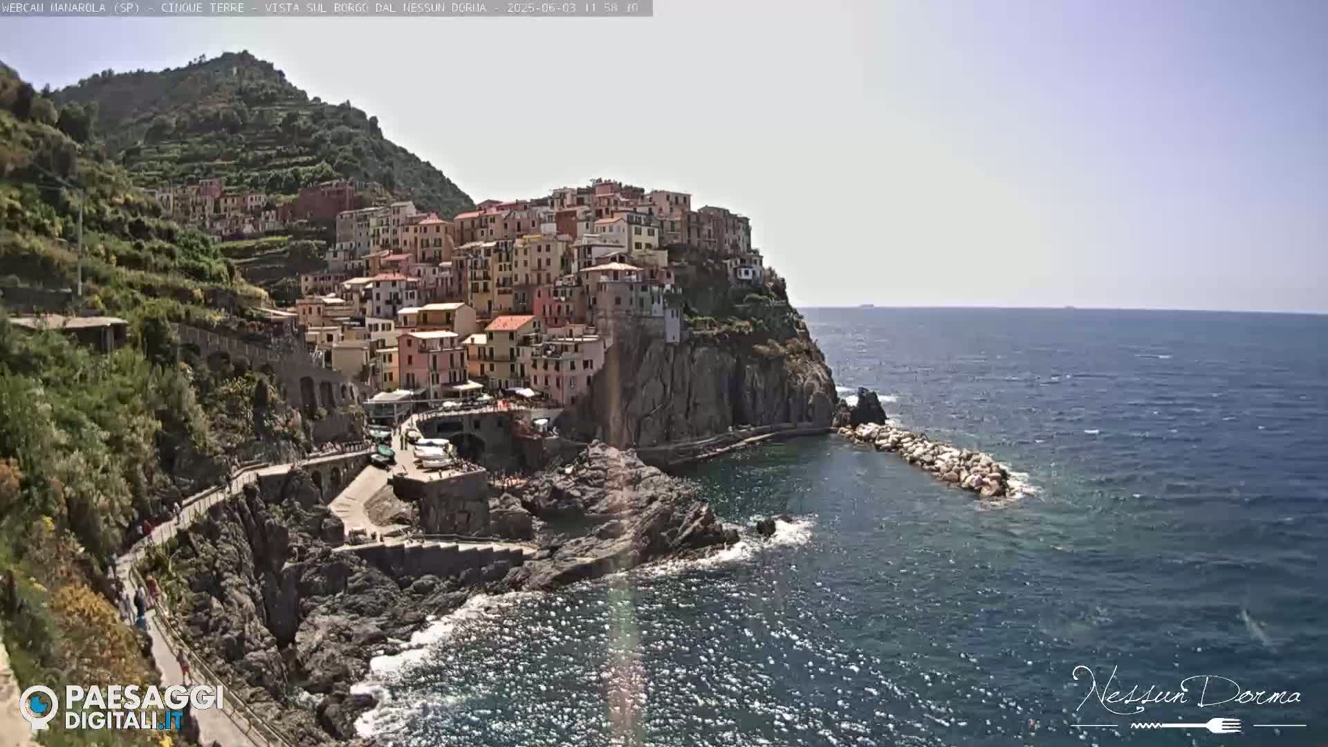 A brightly lit coastal town built on a cliff overlooking a calm, blue sea on a sunny day.