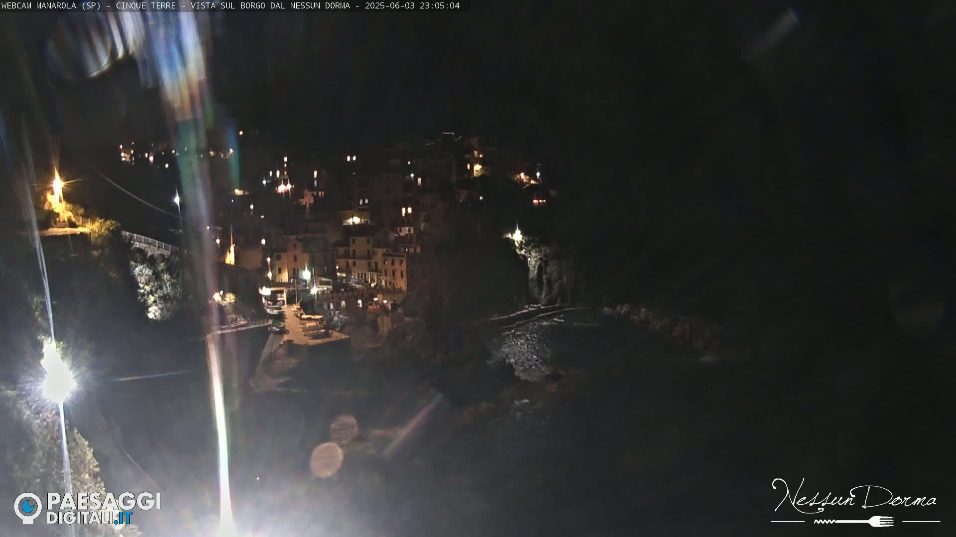 A nighttime view of a coastal village with lights illuminating buildings and a dark body of water below.