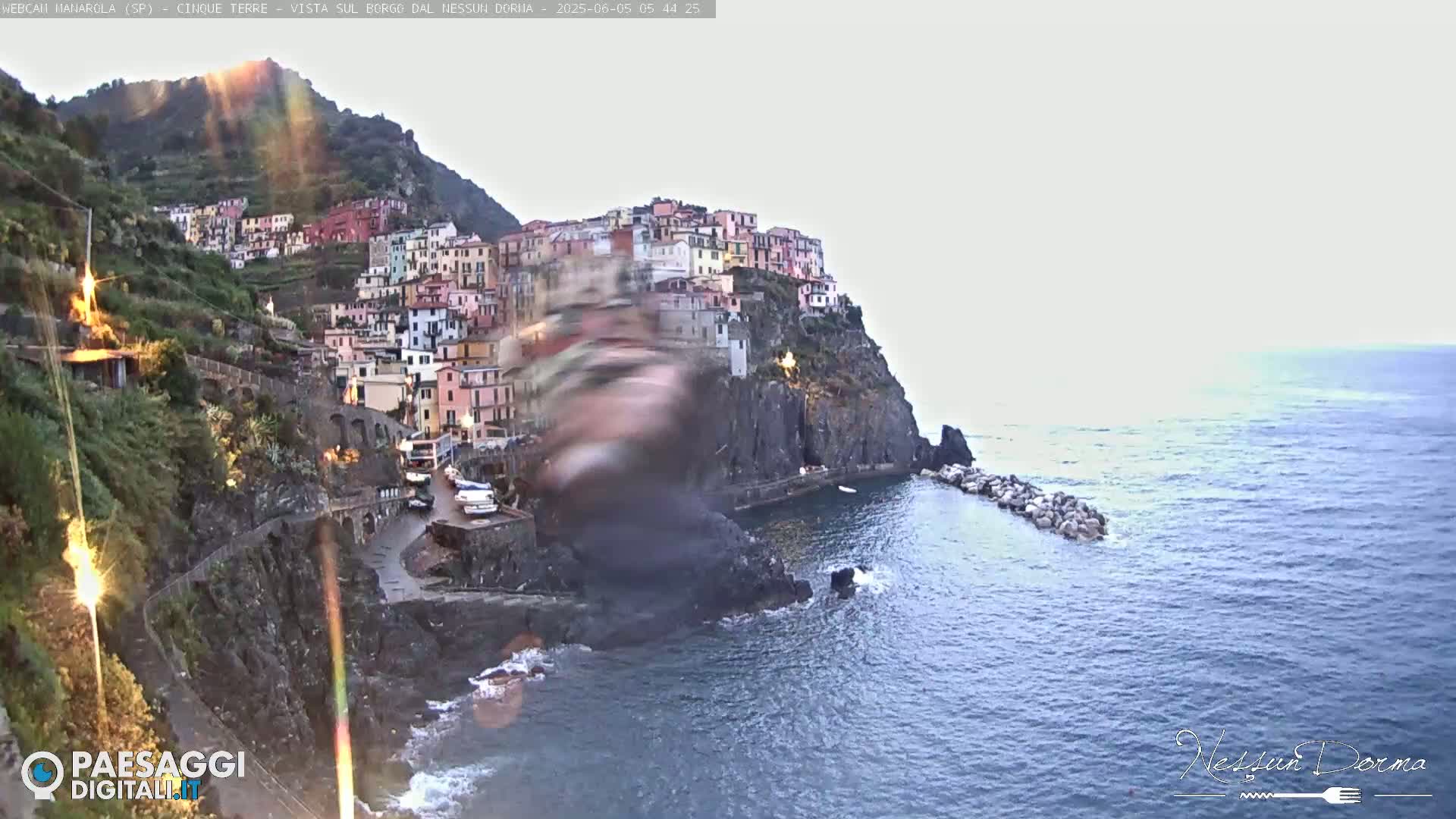 A colorful village clings to a steep cliffside overlooking a calm sea under a mostly clear sky.