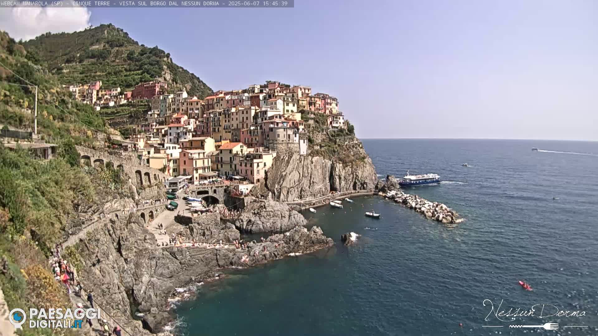 A colorful village clings to a steep cliffside overlooking a calm sea on a sunny day.