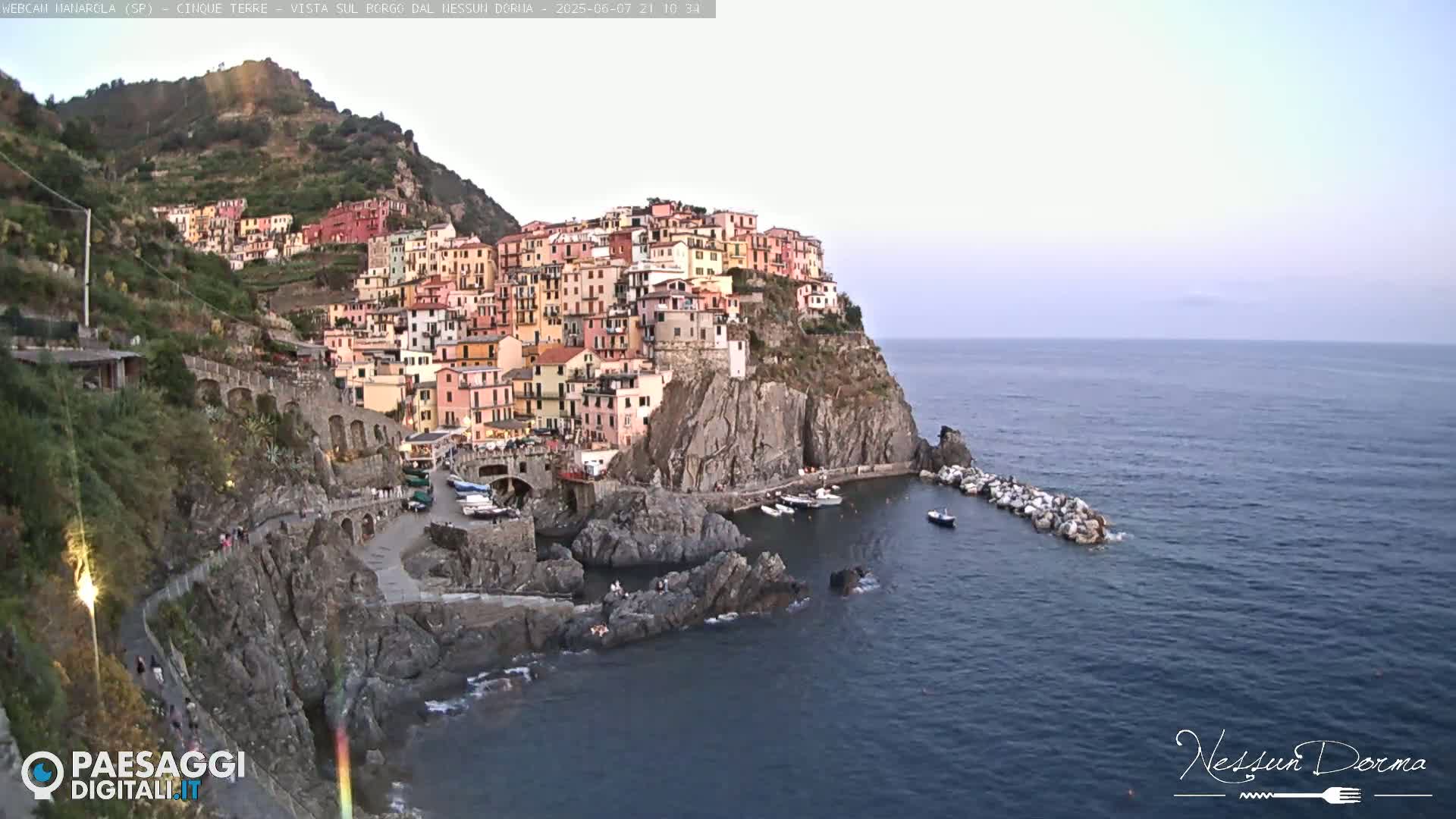 A colorful village clings to a steep cliffside overlooking a calm sea at dusk.