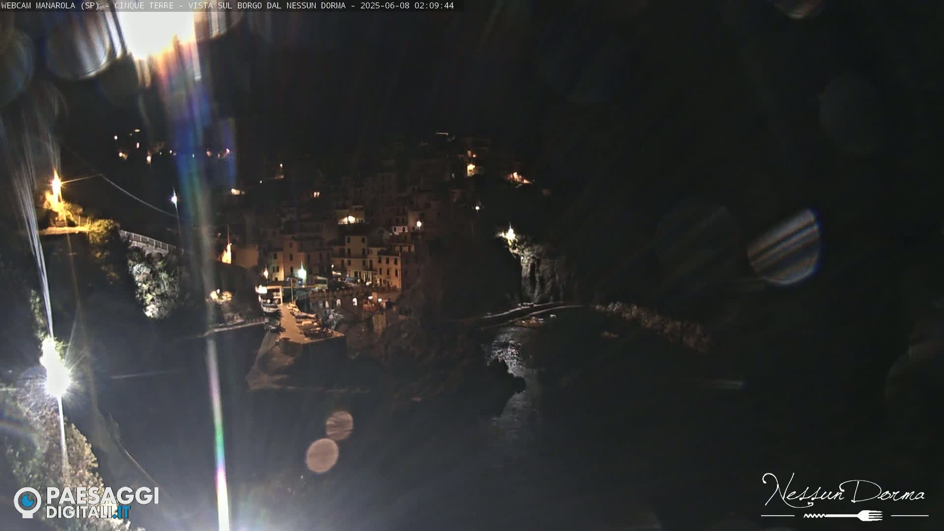 A nighttime, high-angle view of a coastal town nestled on a cliffside, illuminated by scattered lights, with dark water below.