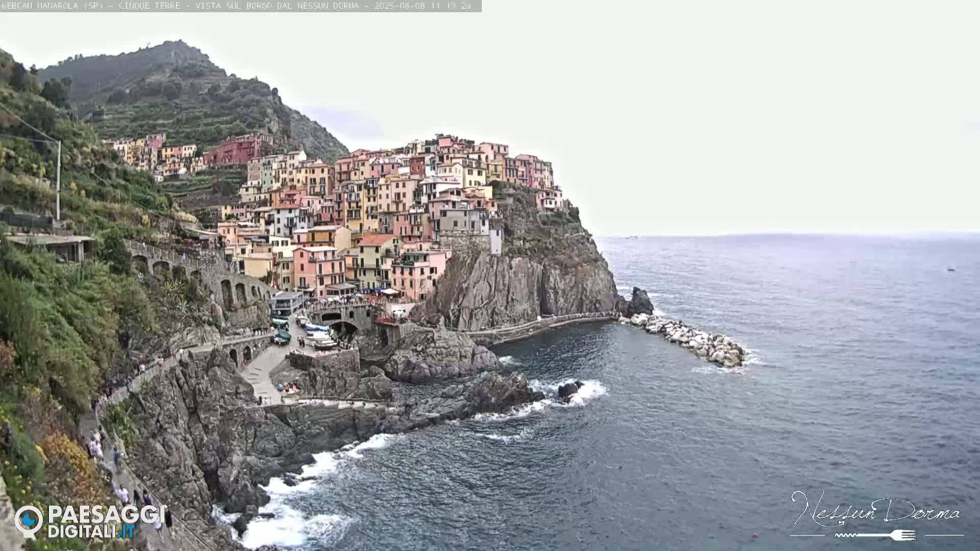 A colorful village clings to a rocky cliffside overlooking a calm sea under an overcast sky.