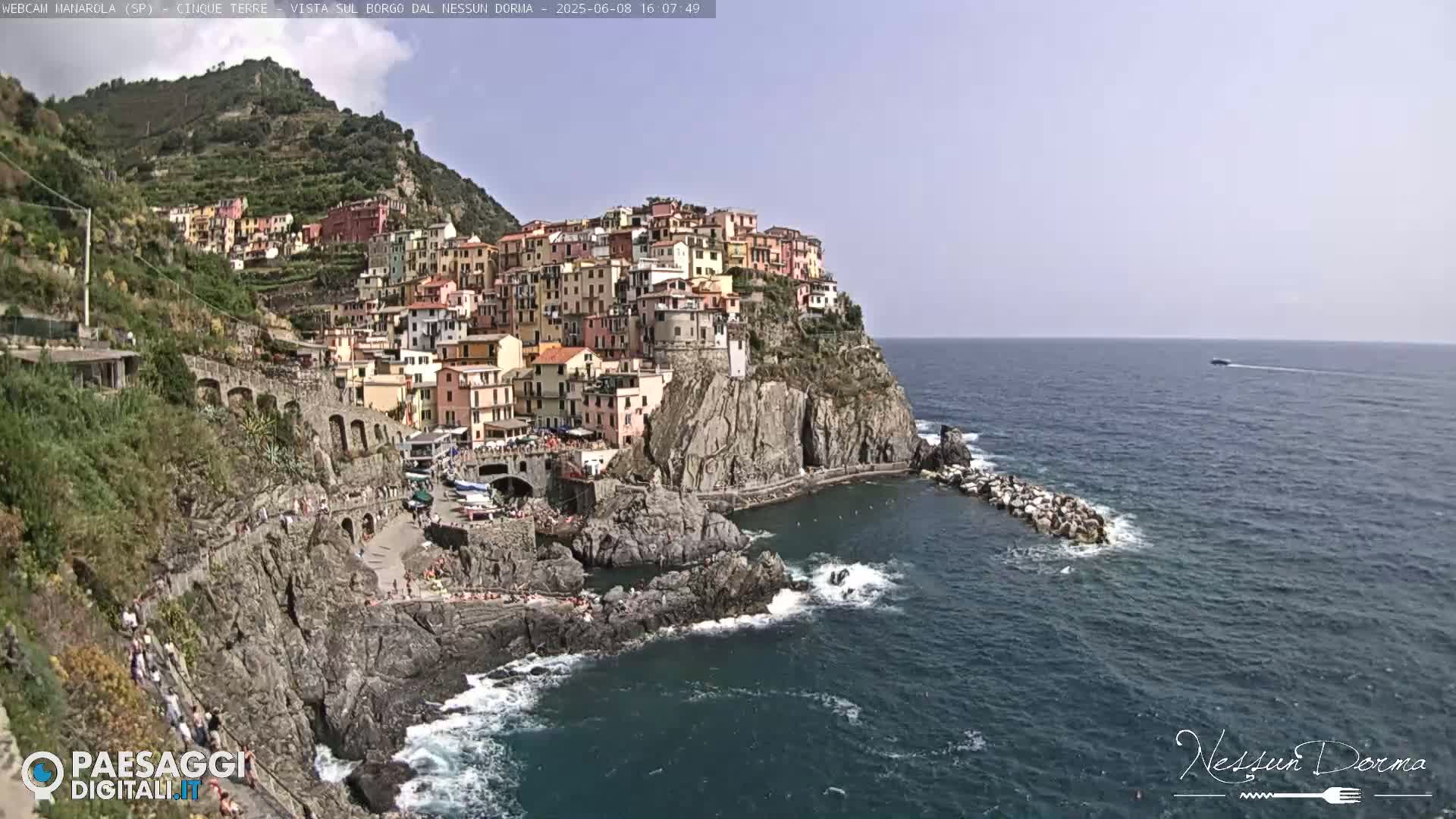 A colorful village clings to a steep cliffside overlooking a calm sea on a sunny day.