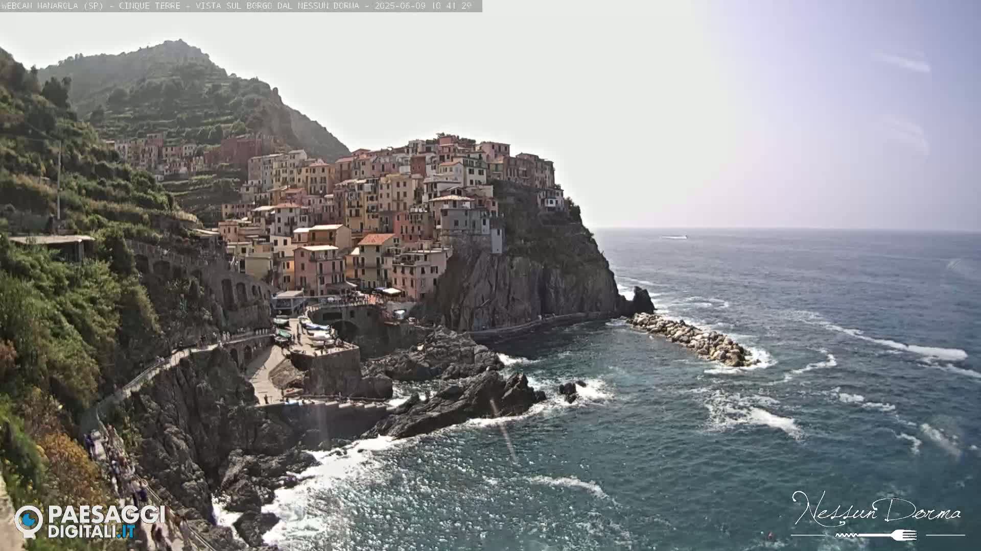 A picturesque coastal village clings to a rocky cliffside overlooking a blue sea under a mostly clear sky.