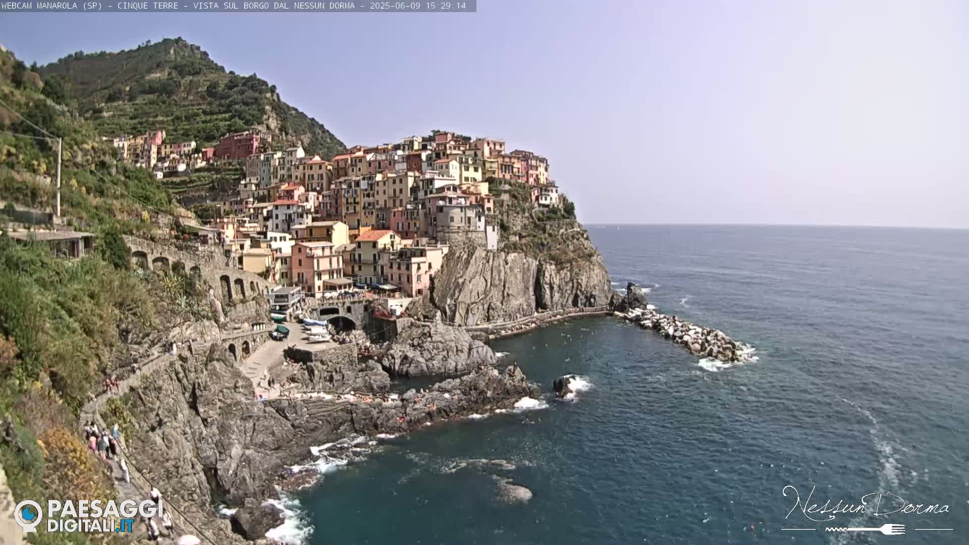 A colorful village clings to a steep, rocky cliffside overlooking a calm, blue sea on a sunny day.