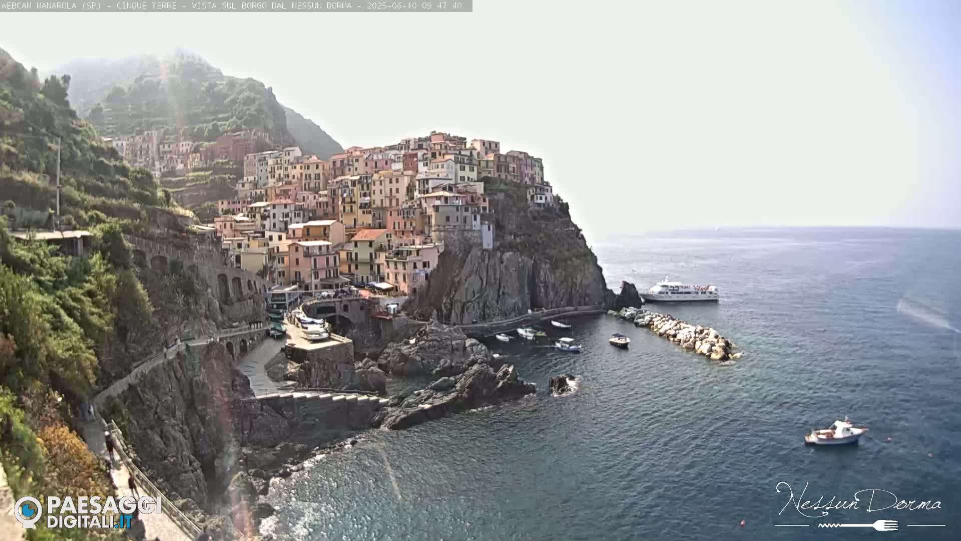 A picturesque coastal village clinging to a steep cliffside overlooks a calm sea with several boats and a larger vessel anchored nearby under a bright, clear sky.