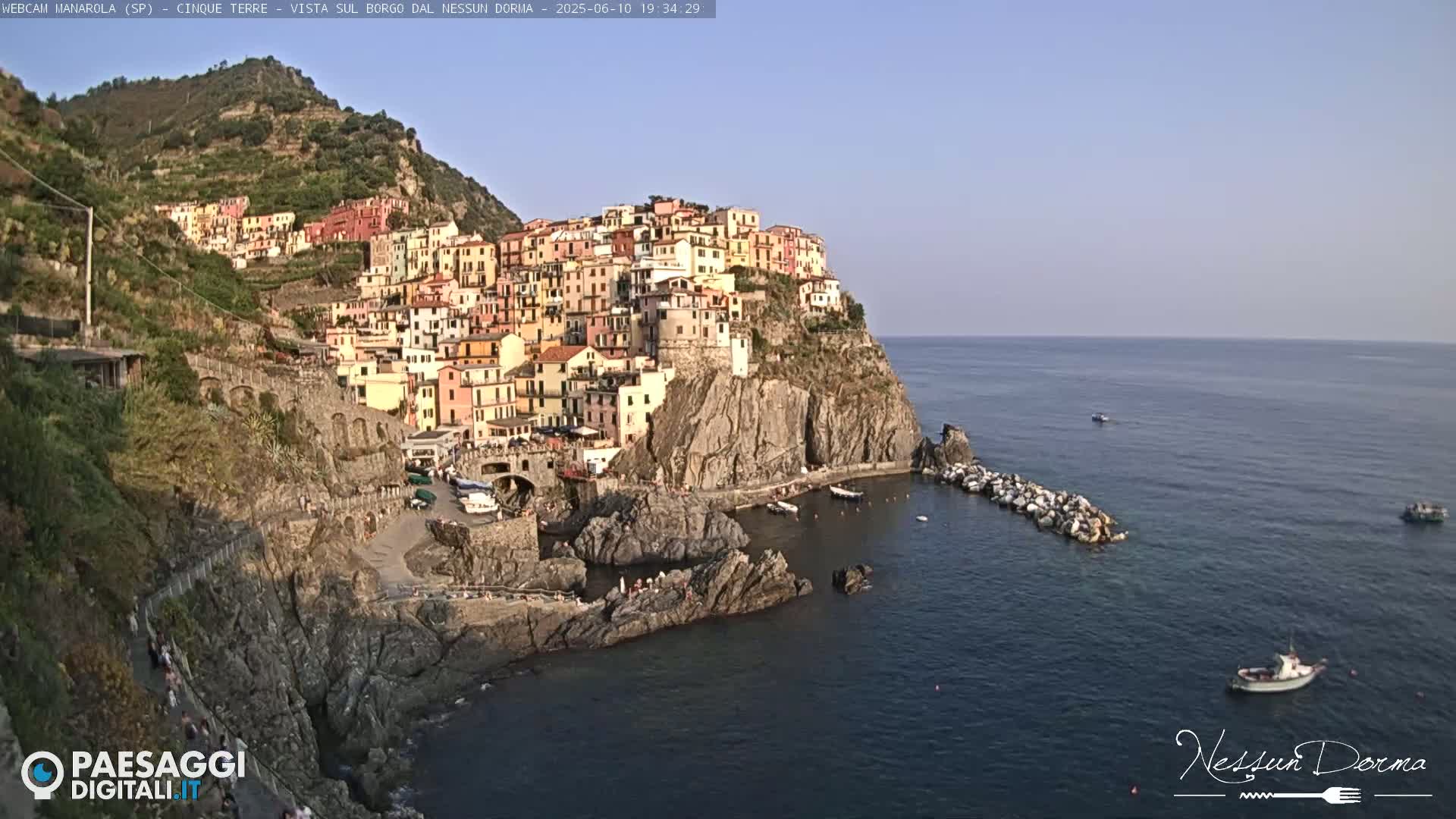 A colorful village clings to a steep cliffside overlooking a calm sea under a clear sky.