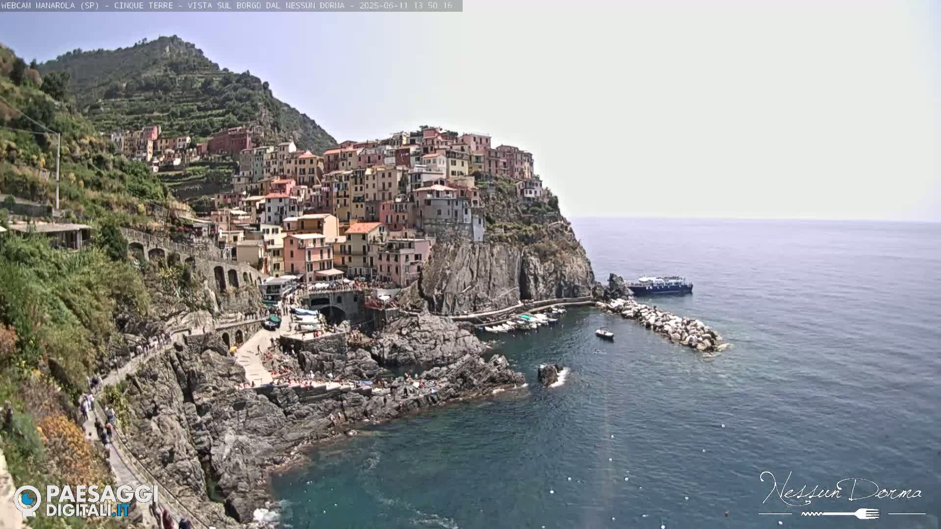 A colorful village clings to a steep cliffside overlooking a calm sea on a bright, sunny day.