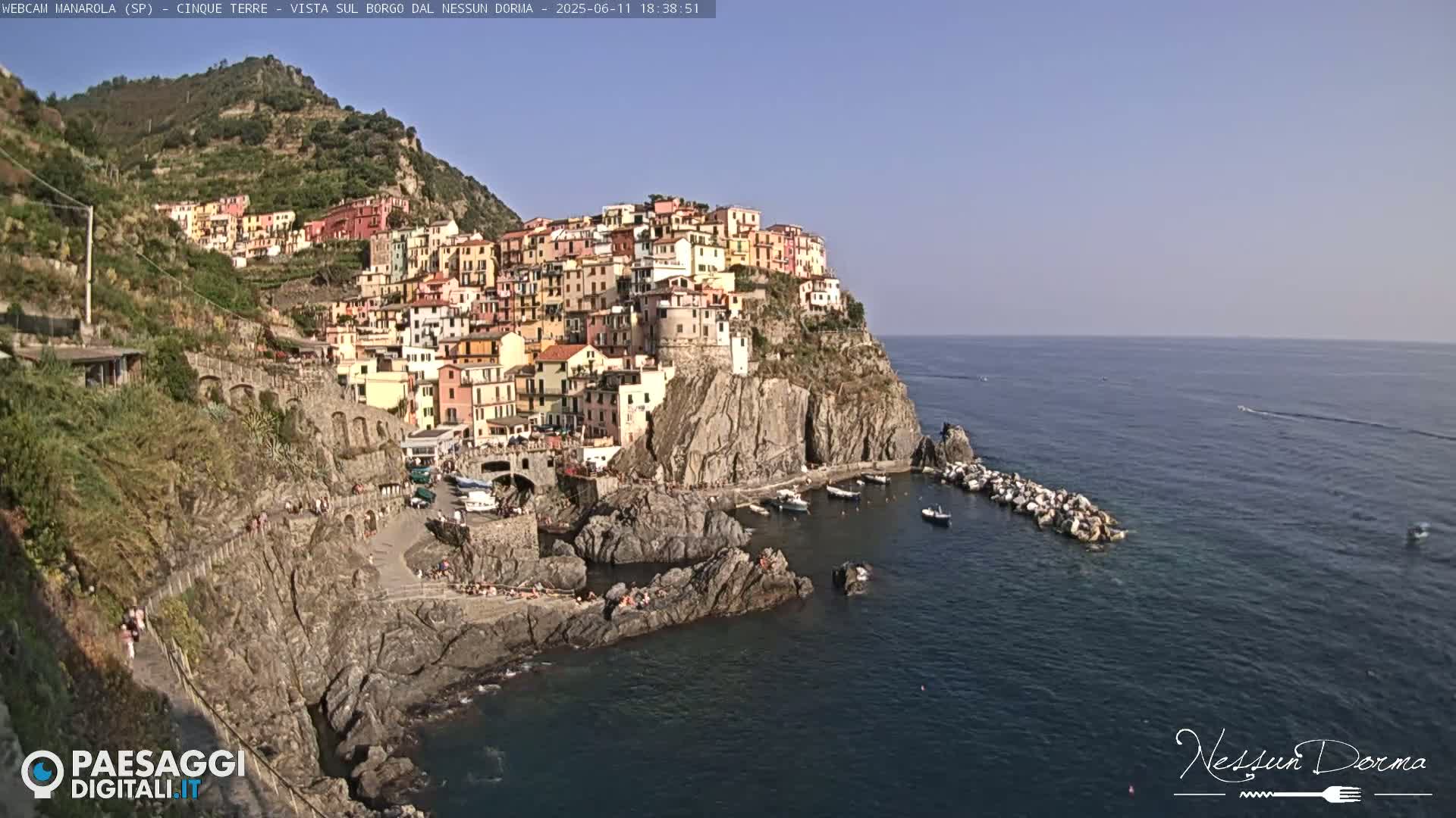 A colorful village clings to a steep cliffside overlooking a calm sea on a sunny day.