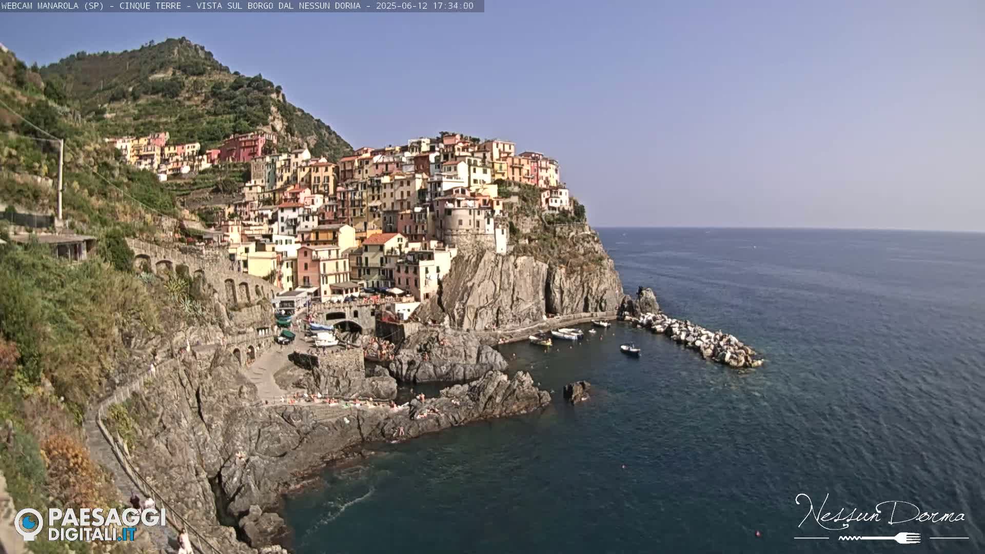 A colorful village clings to a rocky cliffside overlooking a calm, blue sea on a sunny day.