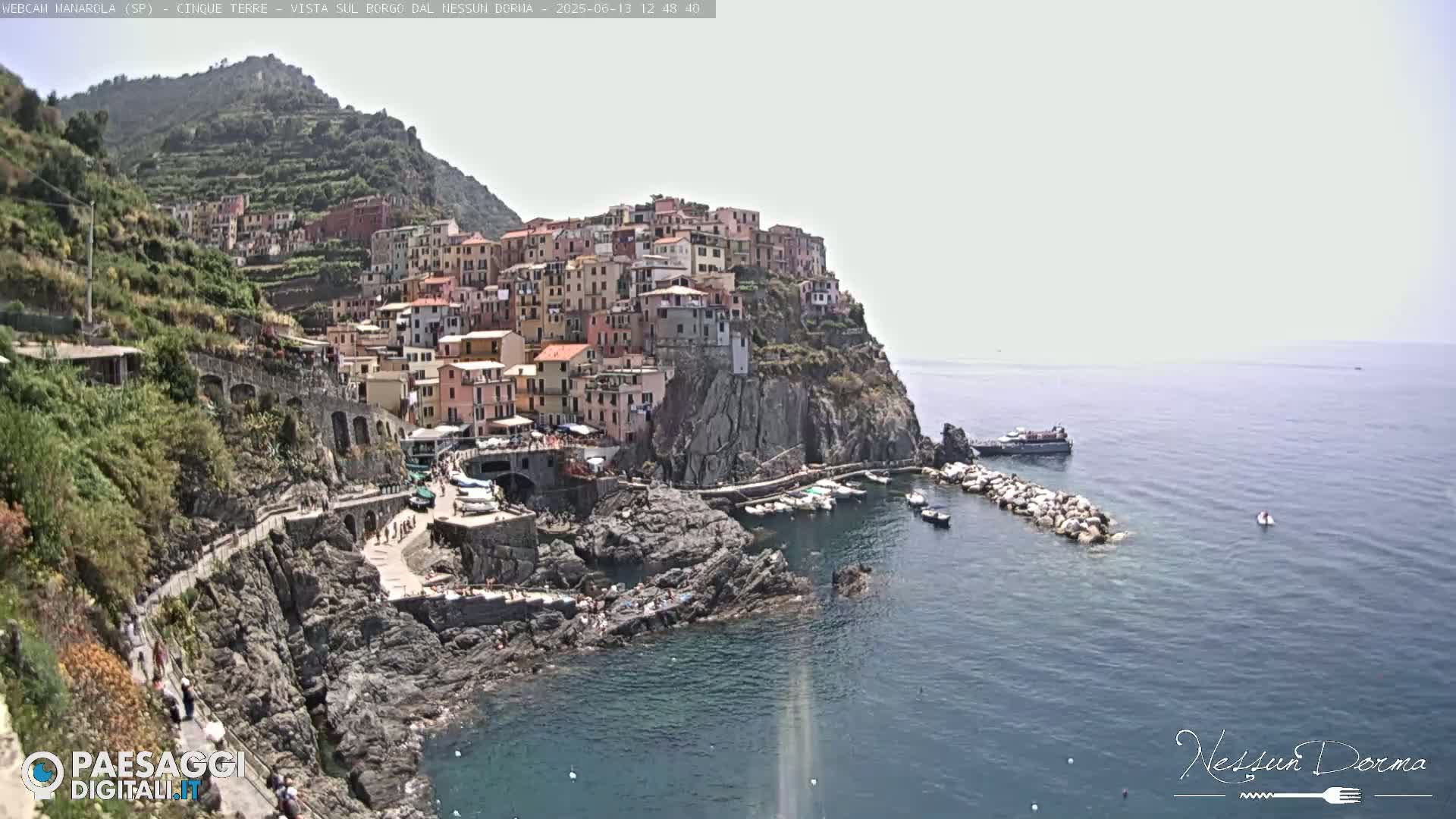A picturesque coastal village clings to a rocky cliffside overlooking a calm sea under a bright, hazy sky.