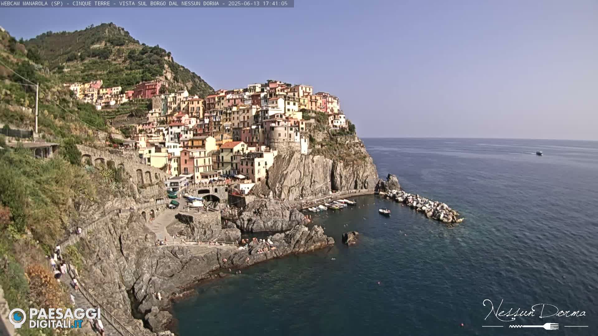 A colorful village clings to a steep cliffside overlooking a calm, blue sea on a sunny day.