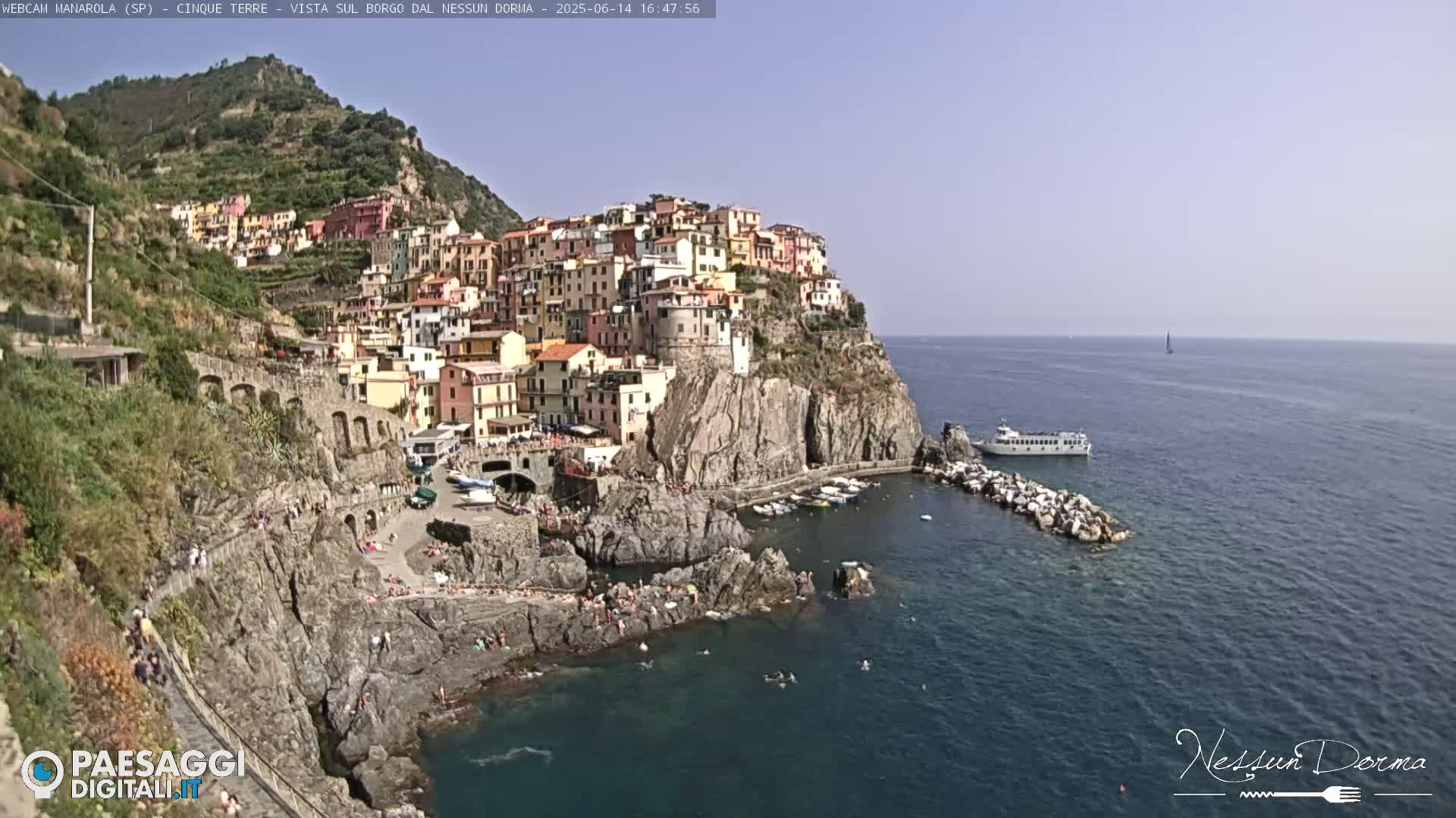 A colorful village clings to a rocky cliffside overlooking a calm, blue sea on a clear, sunny day.