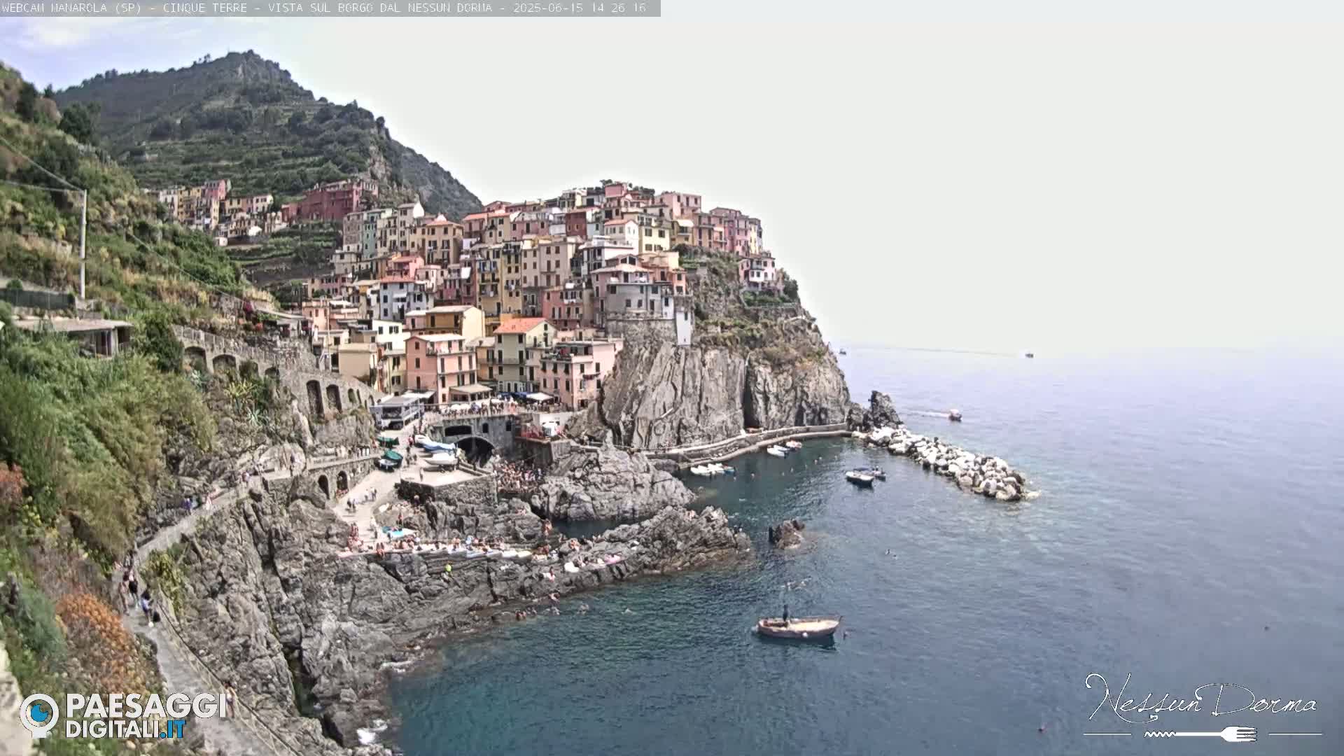 A colorful village clings to a rocky cliffside overlooking a calm sea on a hazy day.